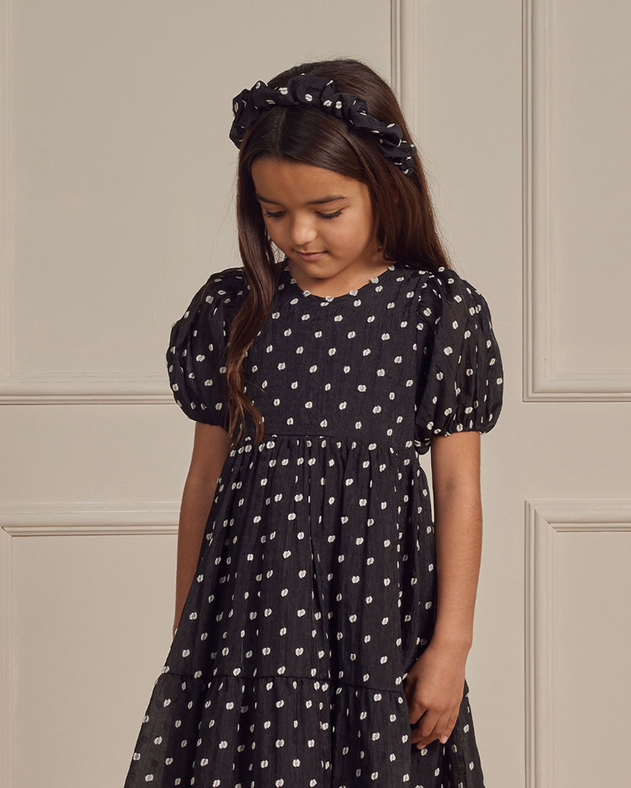 Girl wearing a black polka dot dress and matching headband, looking down with a neutral background.