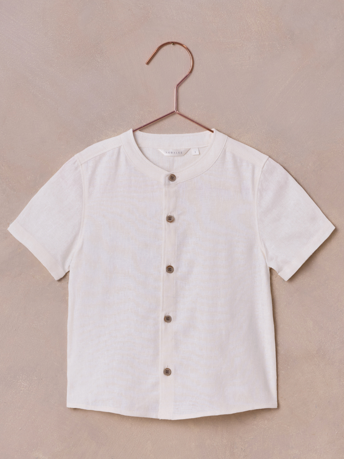 Light beige short-sleeve button-up shirt hanging on a copper-colored hanger against a textured background.