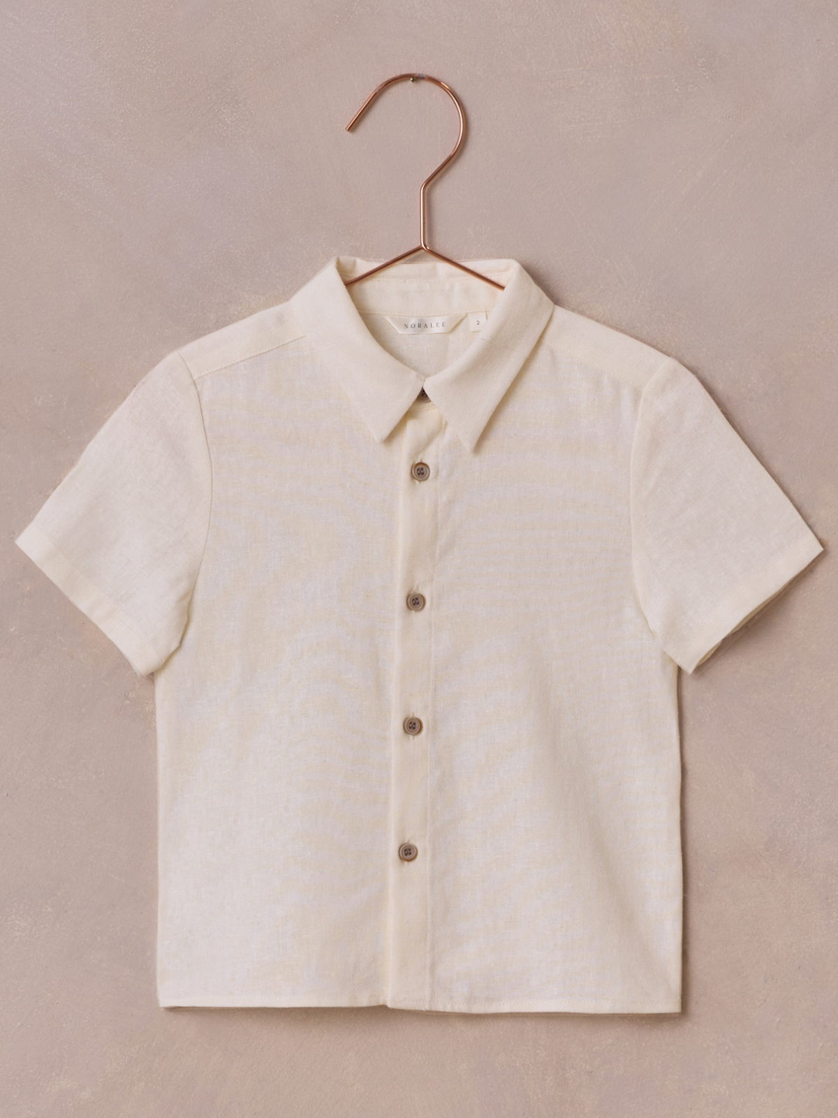 Short-sleeve beige button-up shirt hanging on a copper hook against a textured background.