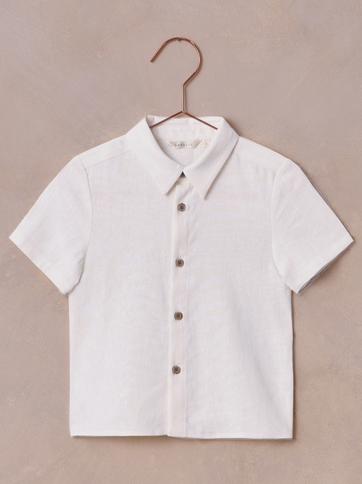 Short-sleeve white shirt with a collar and brown buttons, hanging on a copper hook against a beige background.
