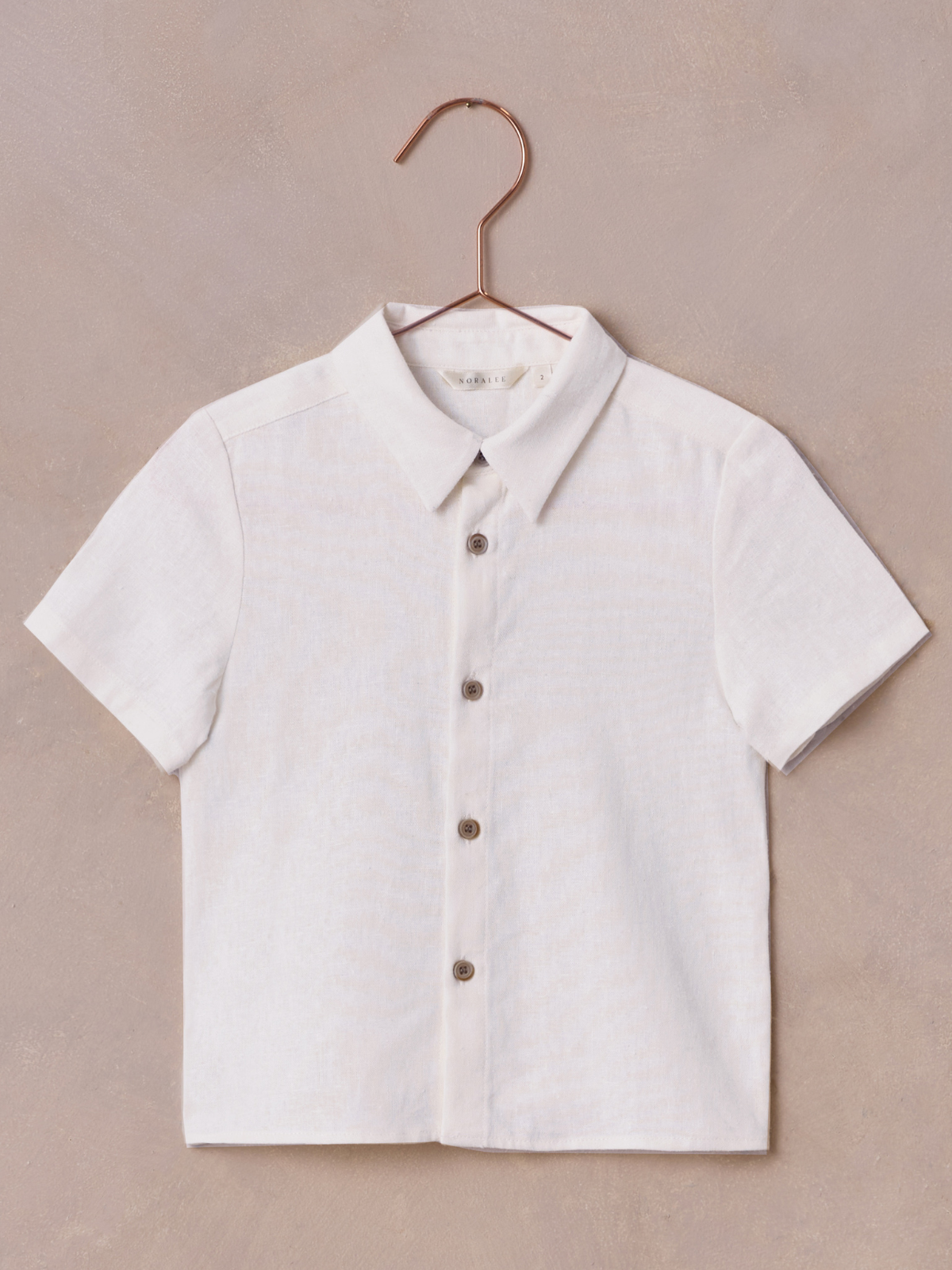 Short-sleeve white shirt with a collar and brown buttons, hanging on a copper hook against a beige background.