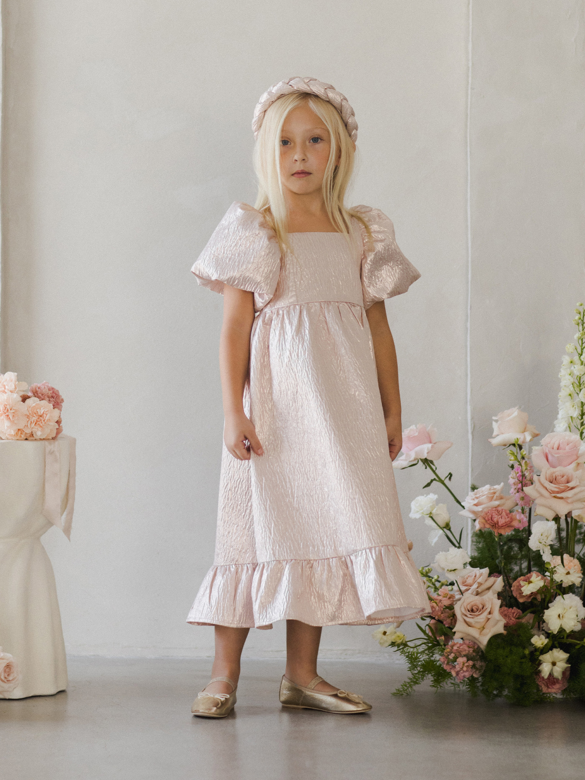 Girl in a pink dress and matching headband, standing near a floral arrangement in a bright indoor setting.