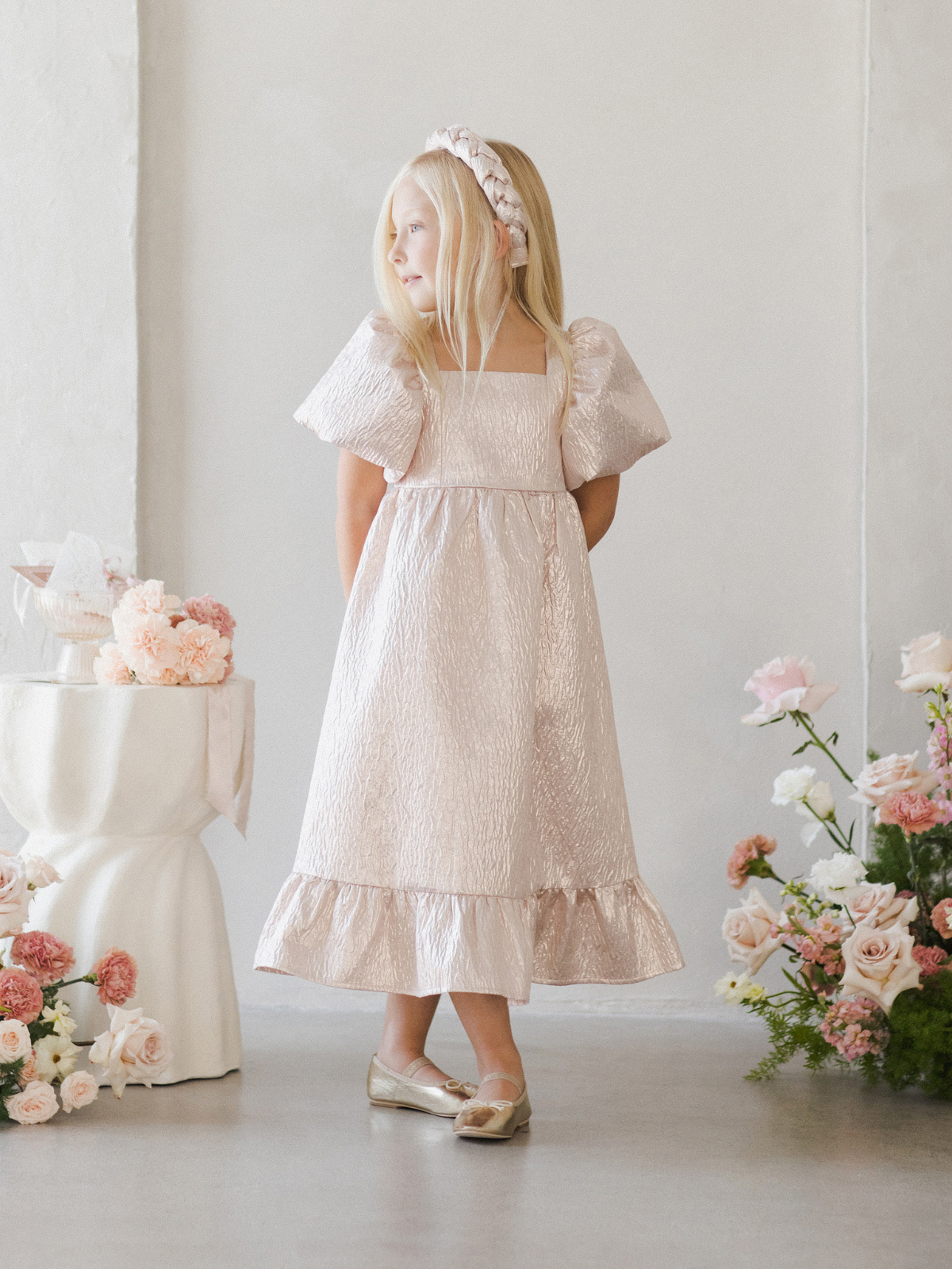 A girl in a pink dress with puffy sleeves stands against a light wall, surrounded by flowers.