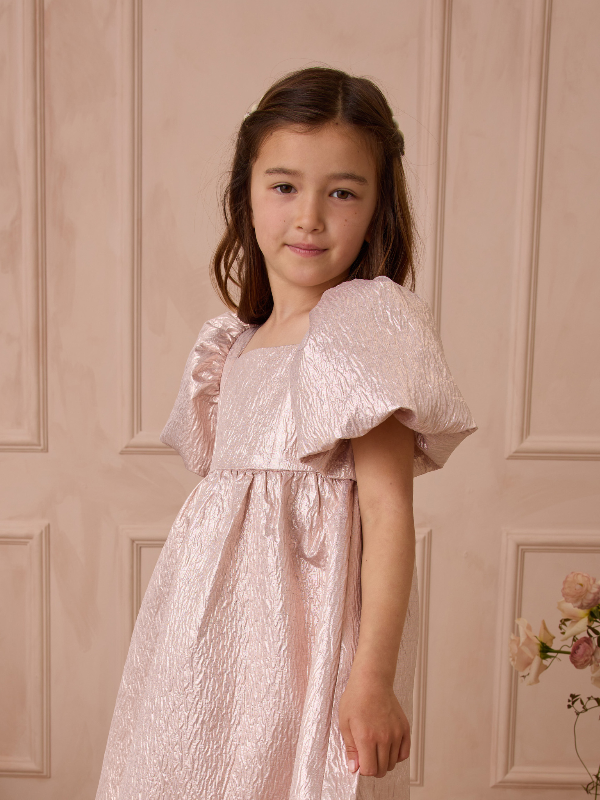 Young girl wearing a shiny pink dress with puff sleeves, standing against a light-colored wall.
