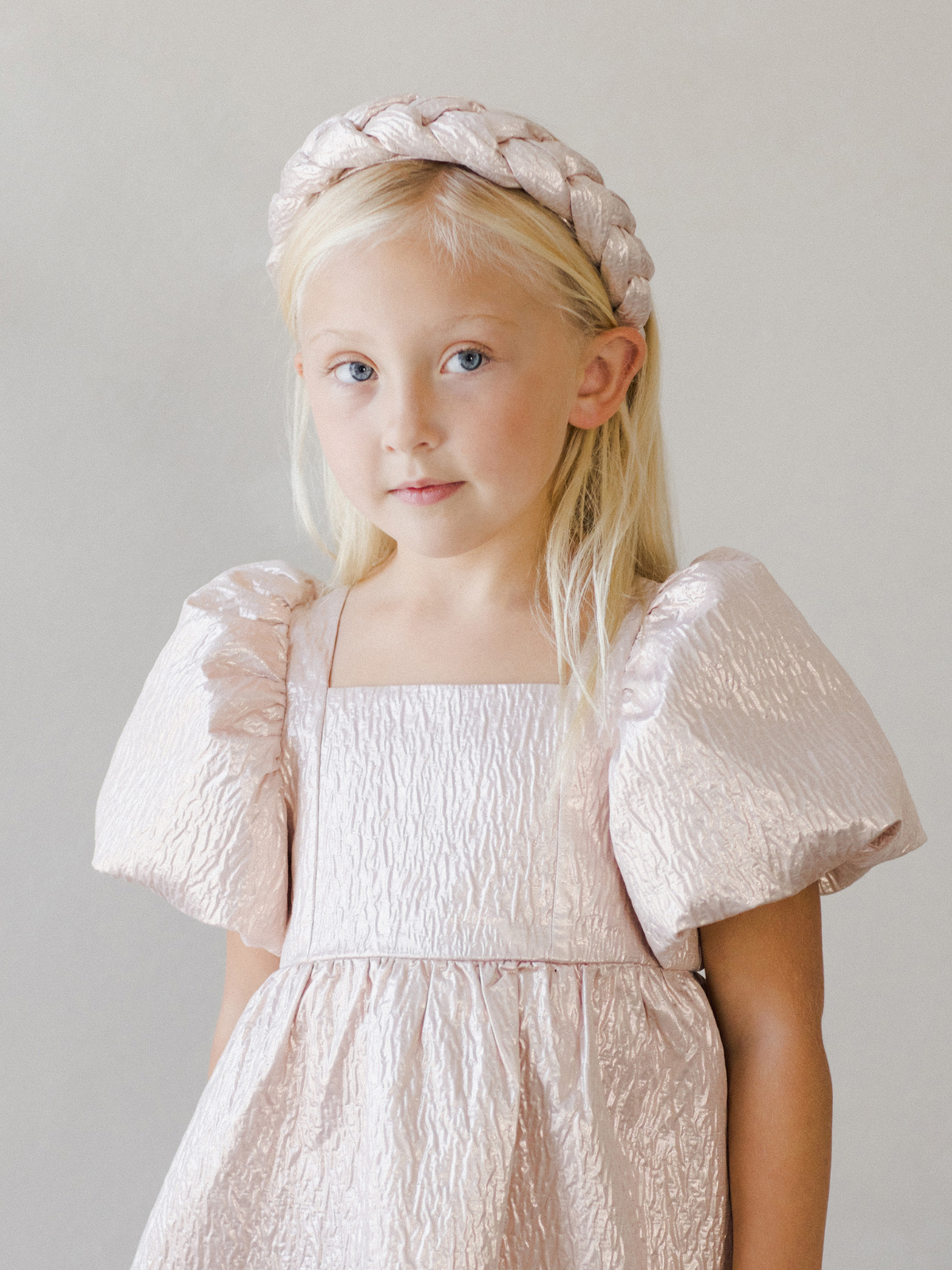 Young girl in a shimmery pink dress with puff sleeves and a braided headband, posing against a soft gray background.