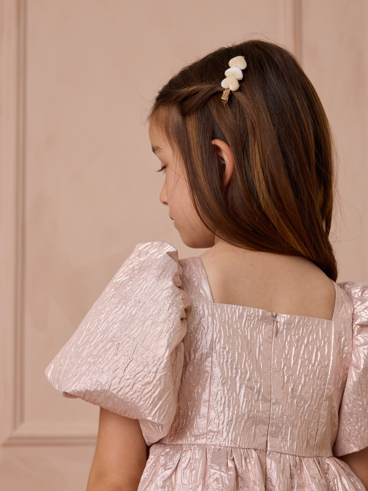 Girl with long hair in a pink dress, facing away, wearing a decorative hair clip, against a textured pink background.