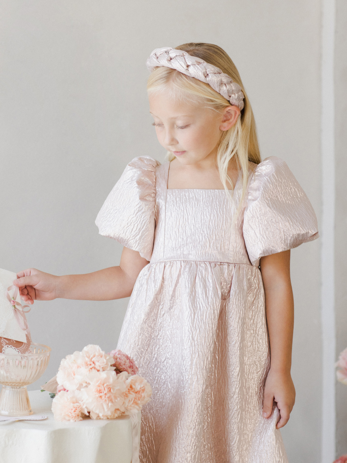 Girl in a pink puffy-sleeve dress holds a vintage bowl, surrounded by flowers and a soft, light background.