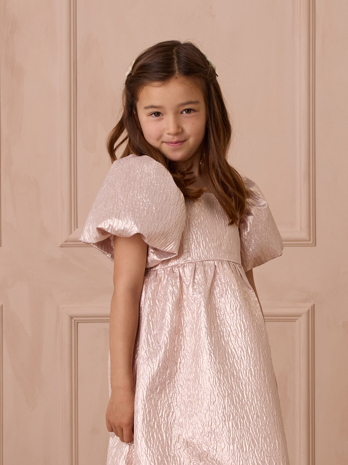 Girl smiling in a shimmering light pink dress with puff sleeves, posed against a soft neutral background.