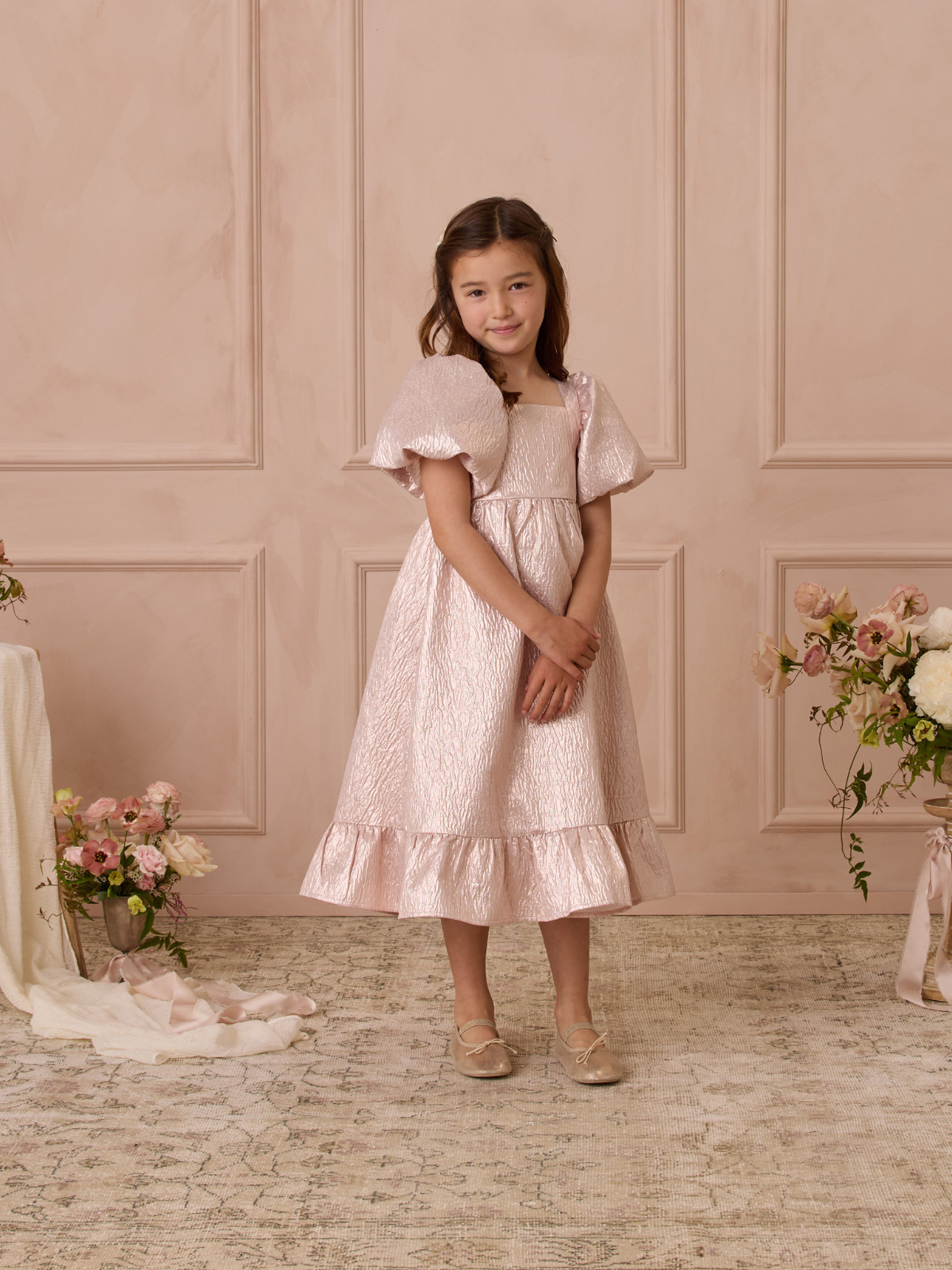 Young girl in a pink dress with puff sleeves, smiling in a softly lit room with floral decorations.