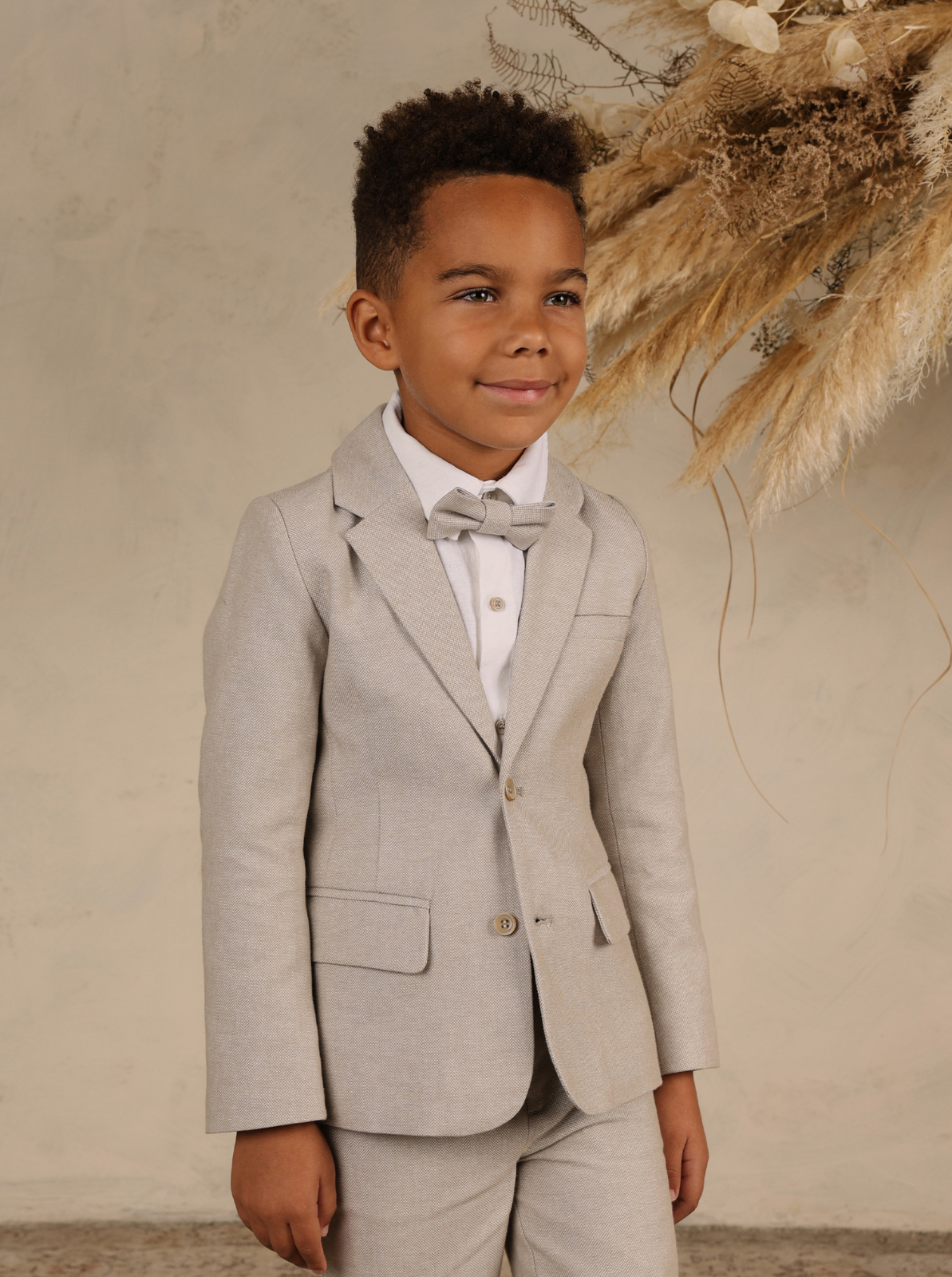 Smiling boy in a light gray suit and bow tie against a neutral background with dried plants.