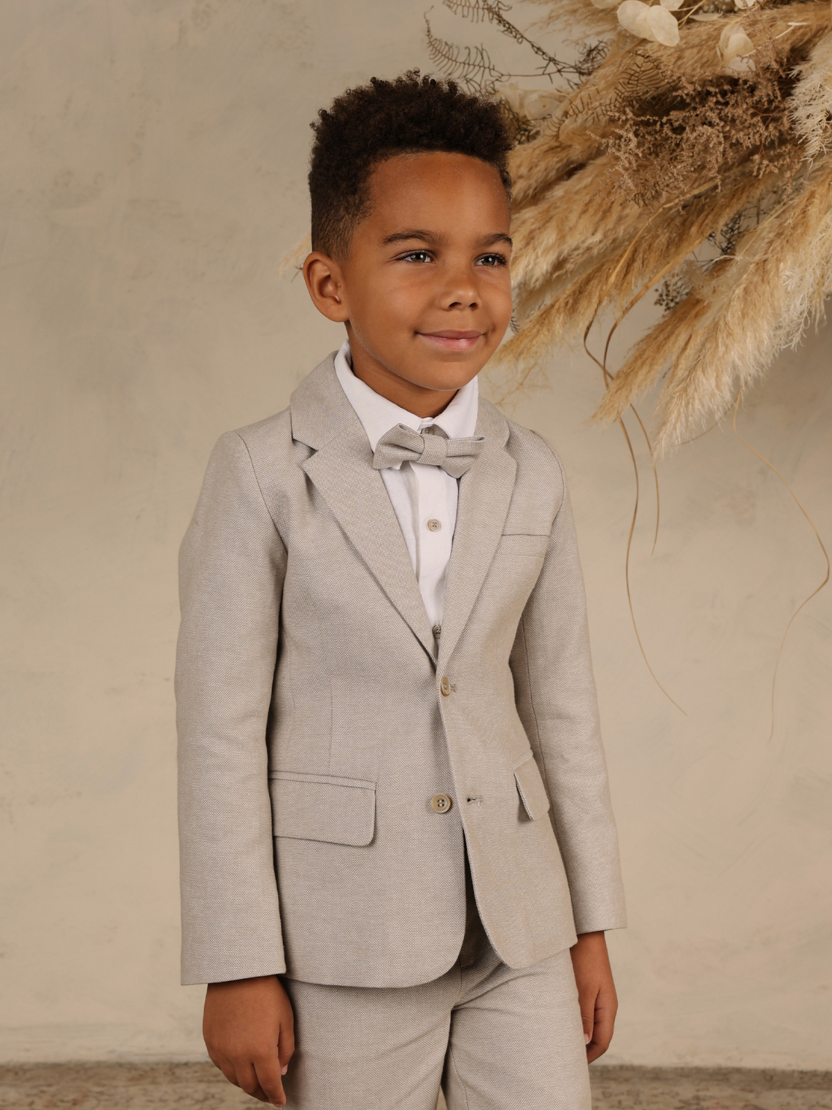 Smiling boy in a light gray suit and bow tie against a neutral background with dried plants.