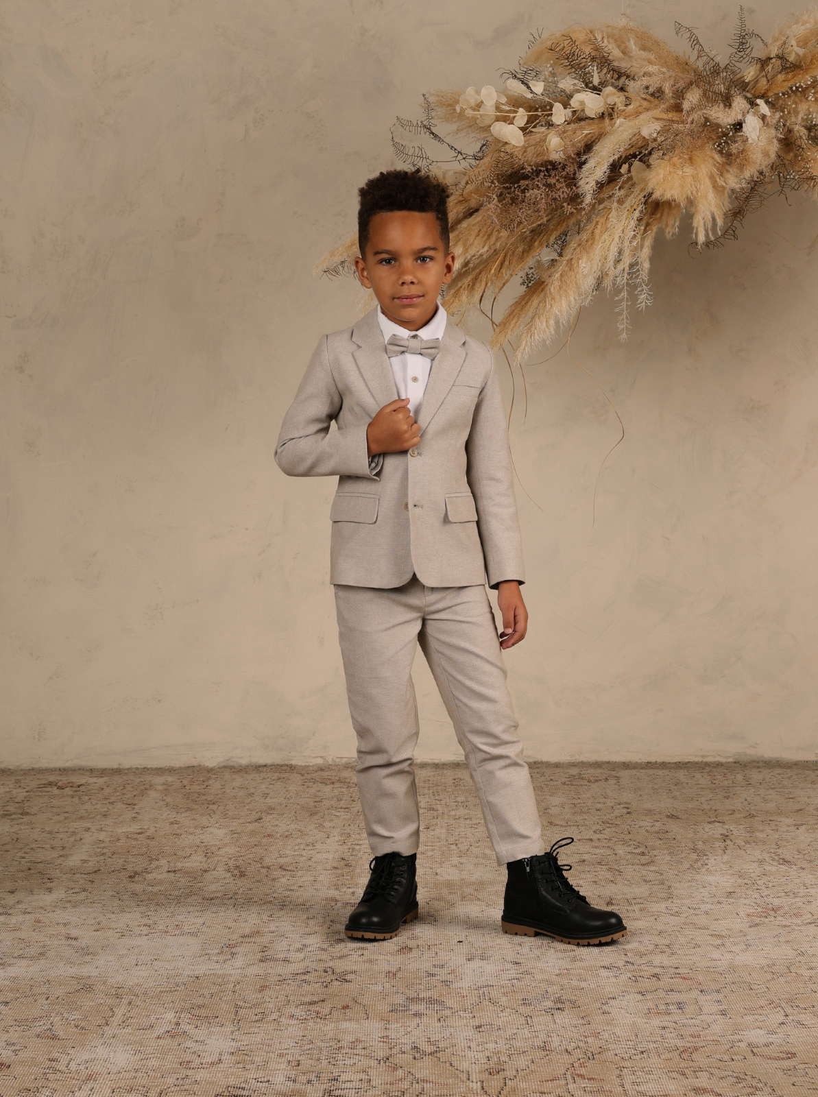 Young boy in a light gray suit and black boots, standing against a textured backdrop with decorative dried flowers.
