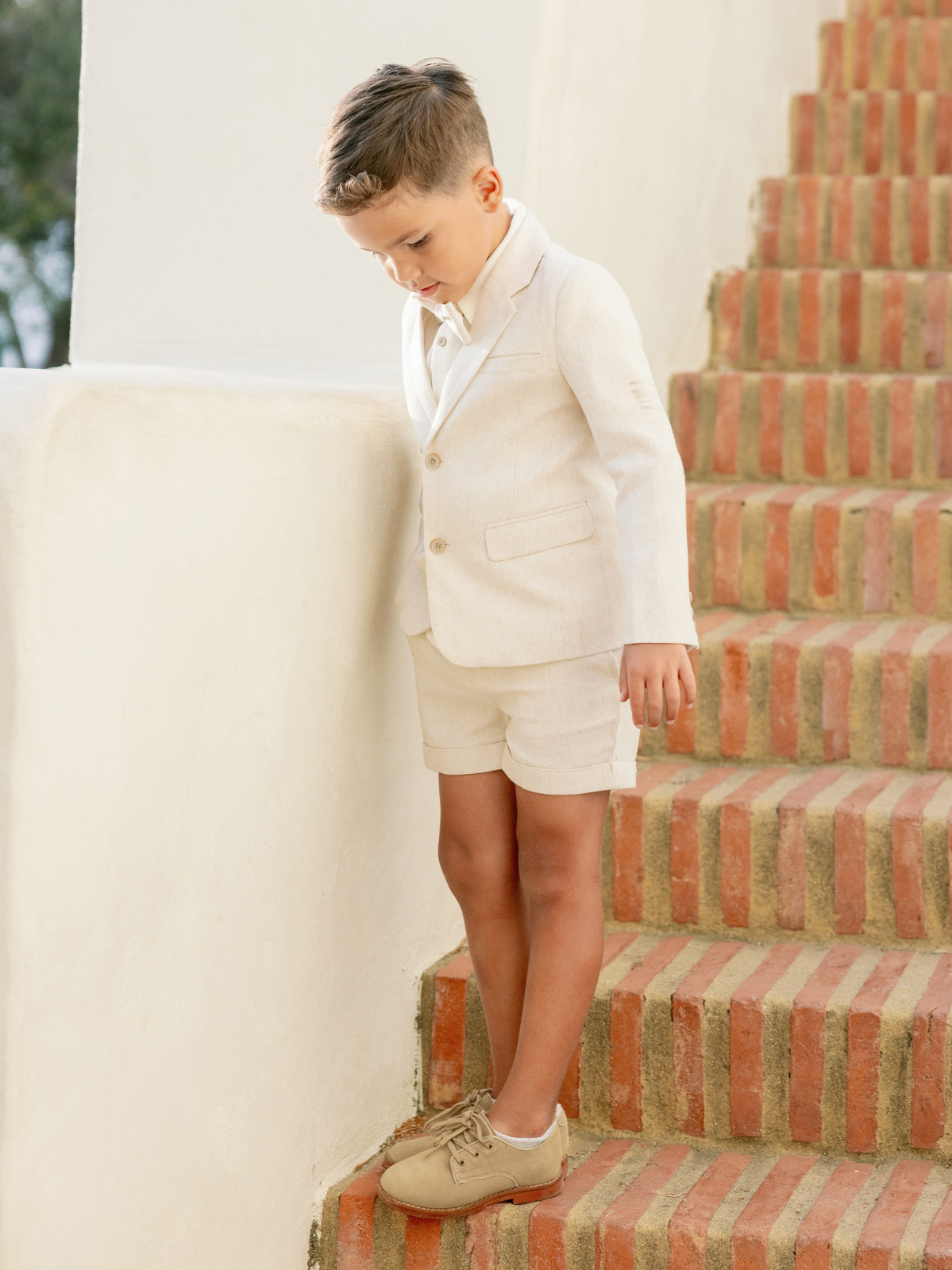 Boy in a light suit and shorts stands on brick stairs, looking down with a thoughtful expression.