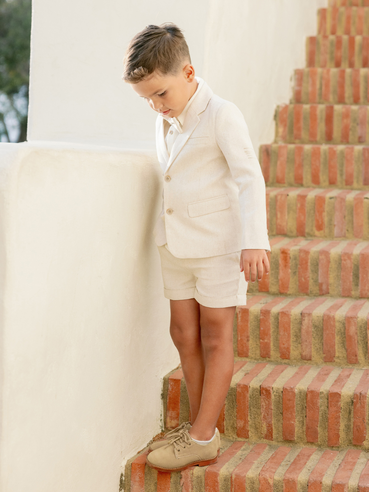 Boy in a light suit and shorts stands on brick stairs, looking down with a thoughtful expression.