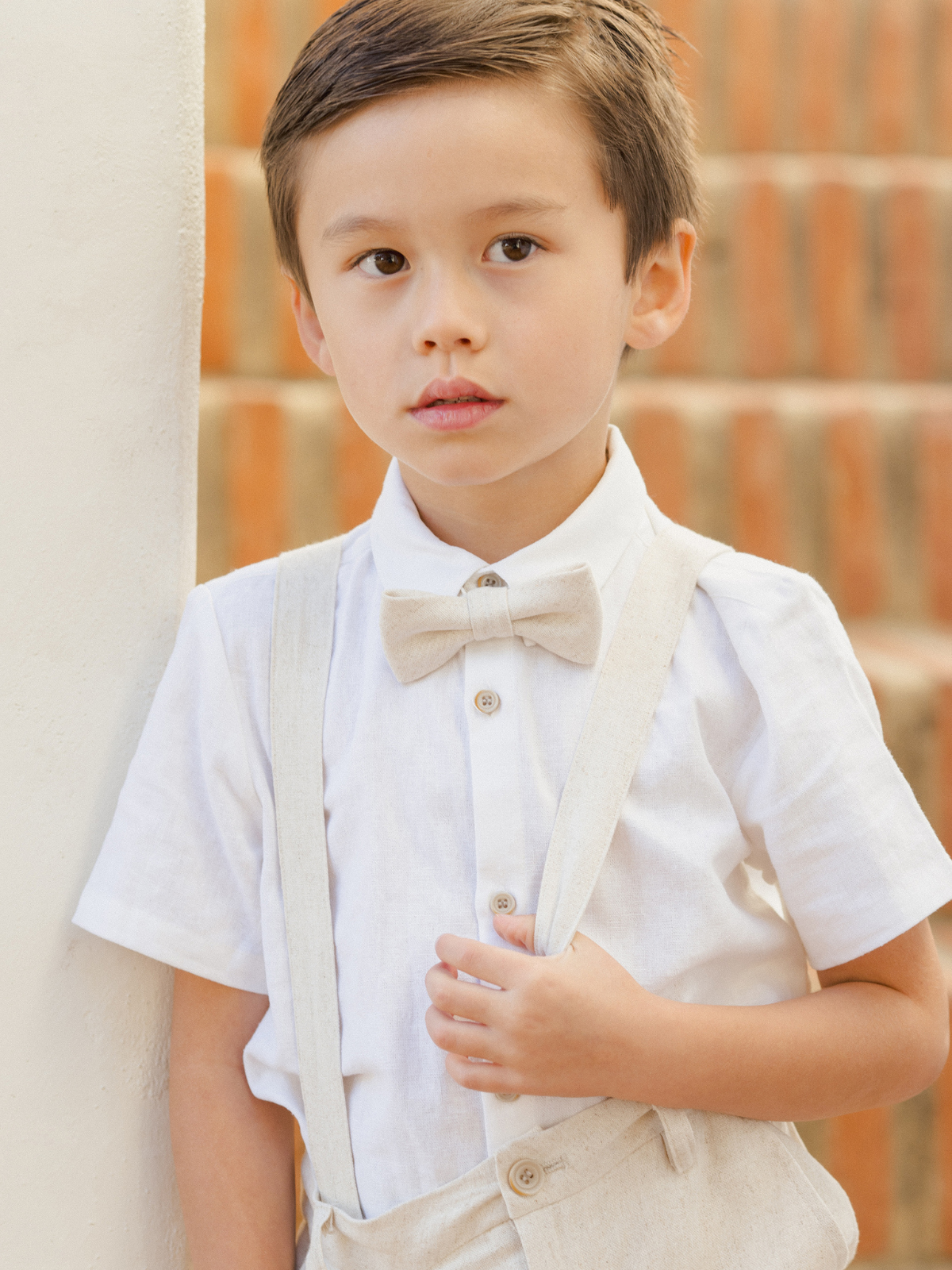 Young boy in a white shirt and bow tie, holding suspenders, standing against a brick wall.