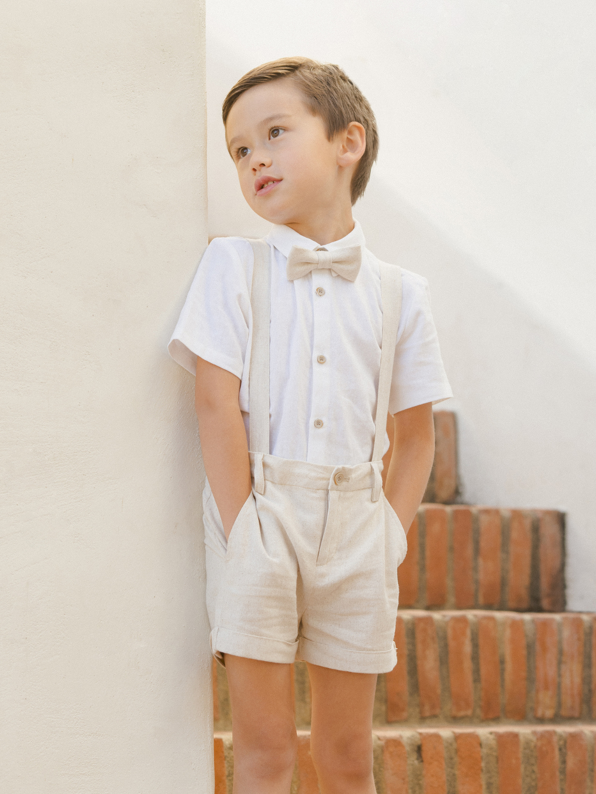 Young boy in a white shirt and bow tie, wearing beige shorts, leaning against a wall near brick steps.