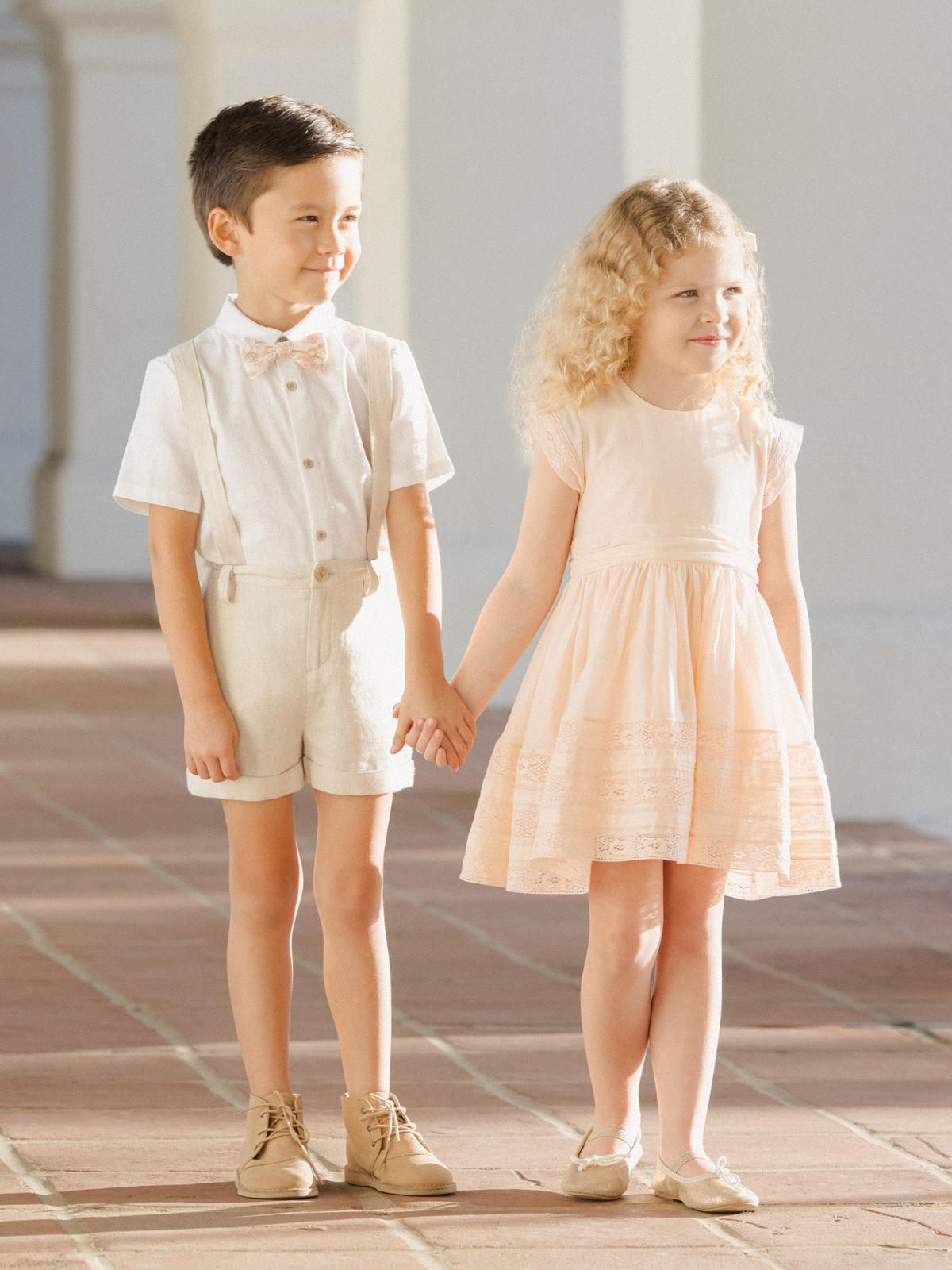 Two smiling children hold hands, dressed in a light outfit and a pastel dress, standing on a sunlit pathway.