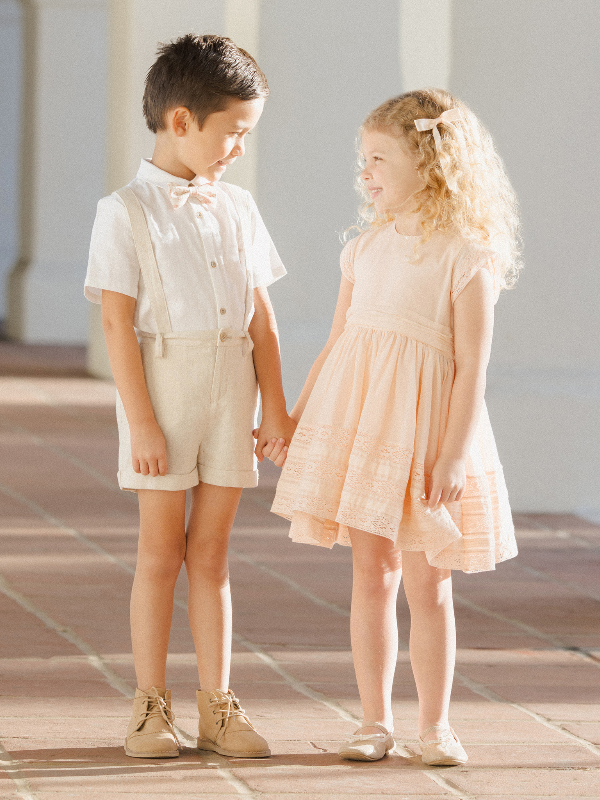 Two children, a boy in a light outfit and a girl in a peach dress, holding hands and smiling in a sunny setting.
