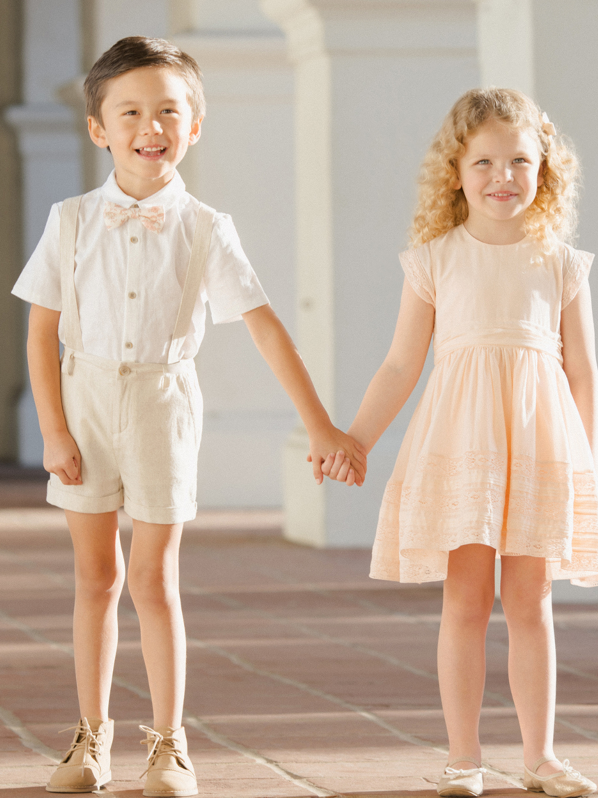 Two children, a boy in a bow tie and shorts and a girl in a dress, hold hands while smiling outdoors.