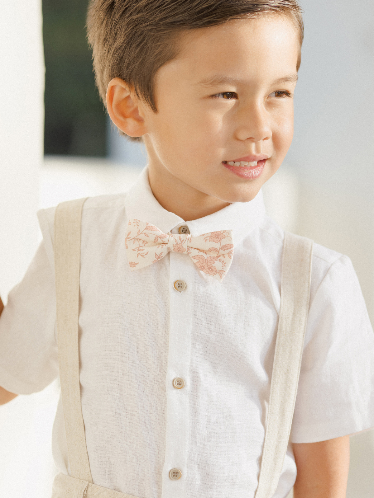 Smiling boy in a white shirt with a floral bow tie and suspenders, standing against a light background.