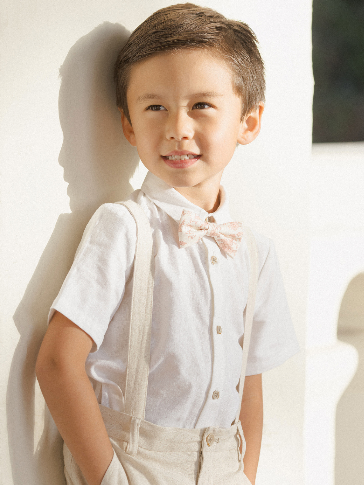 Smiling young boy in a white shirt and bow tie, leaning against a wall, showcasing a playful expression.