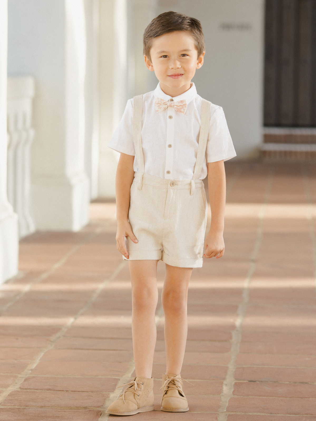Young boy in a stylish outfit with suspenders, posing indoors with natural light on a tiled floor.
