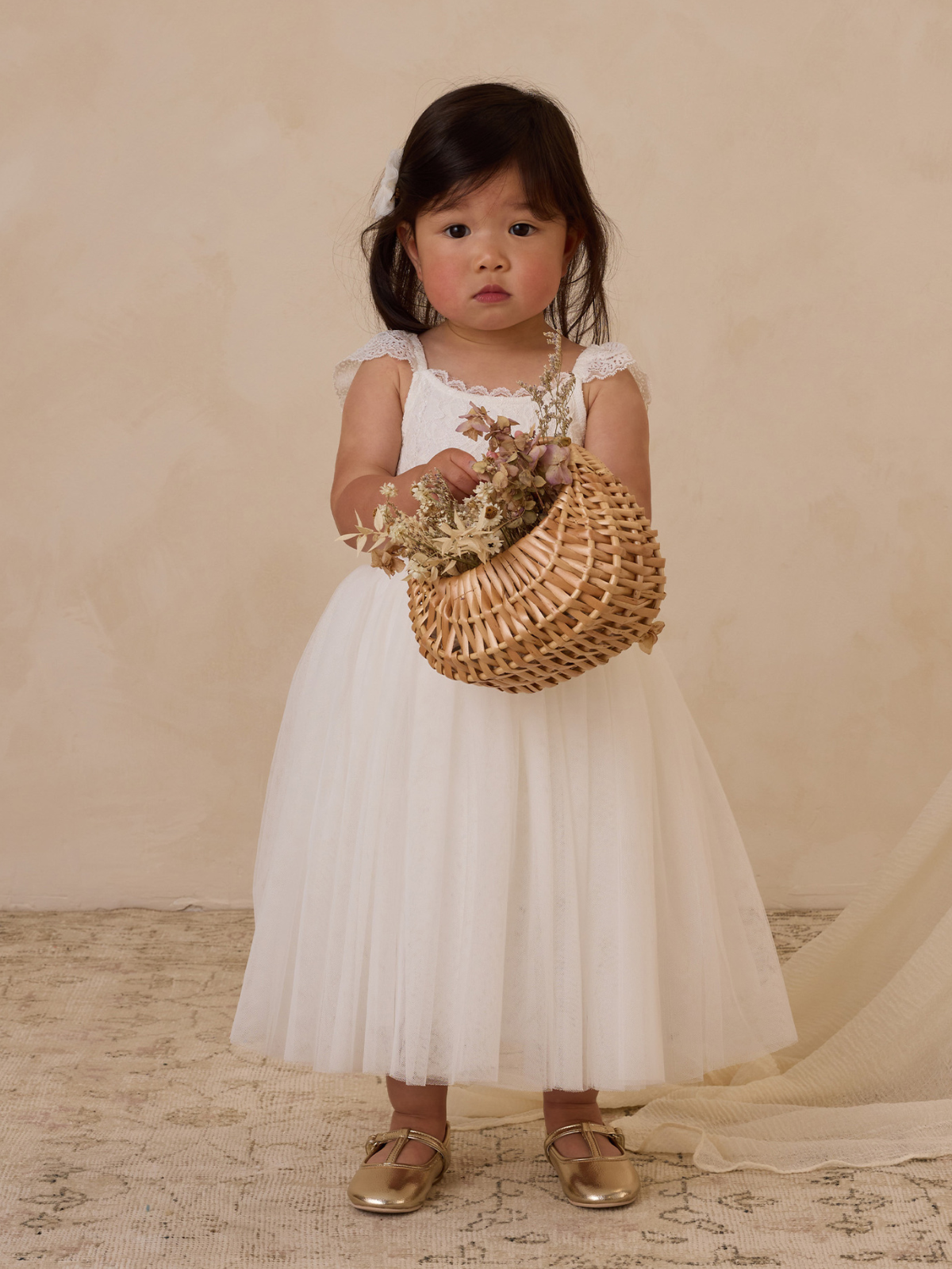Young girl in a white dress holding a woven basket with dried flowers, standing on a textured rug against a light backdrop.