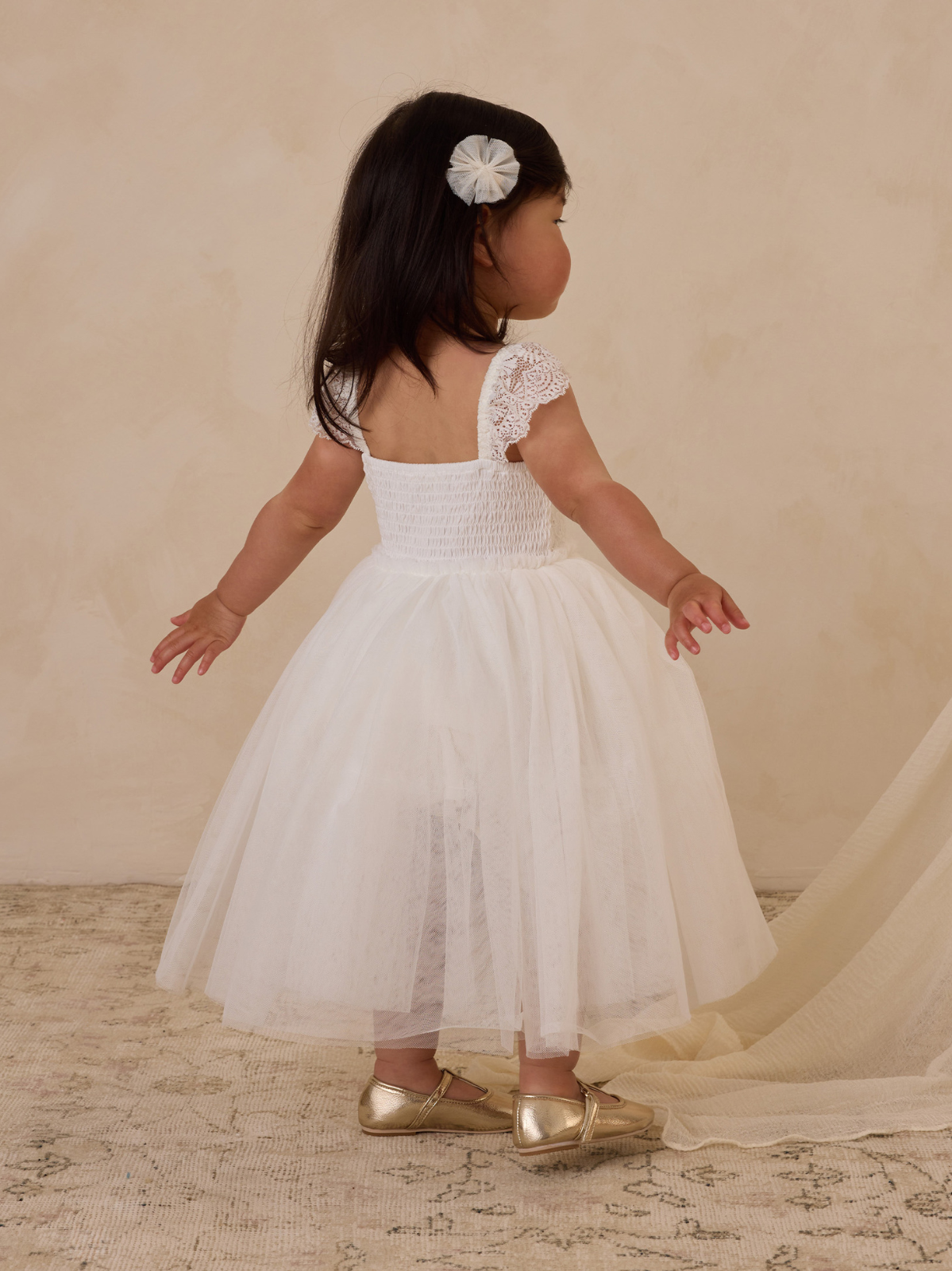 A toddler in a white dress and golden shoes, facing away against a neutral background.