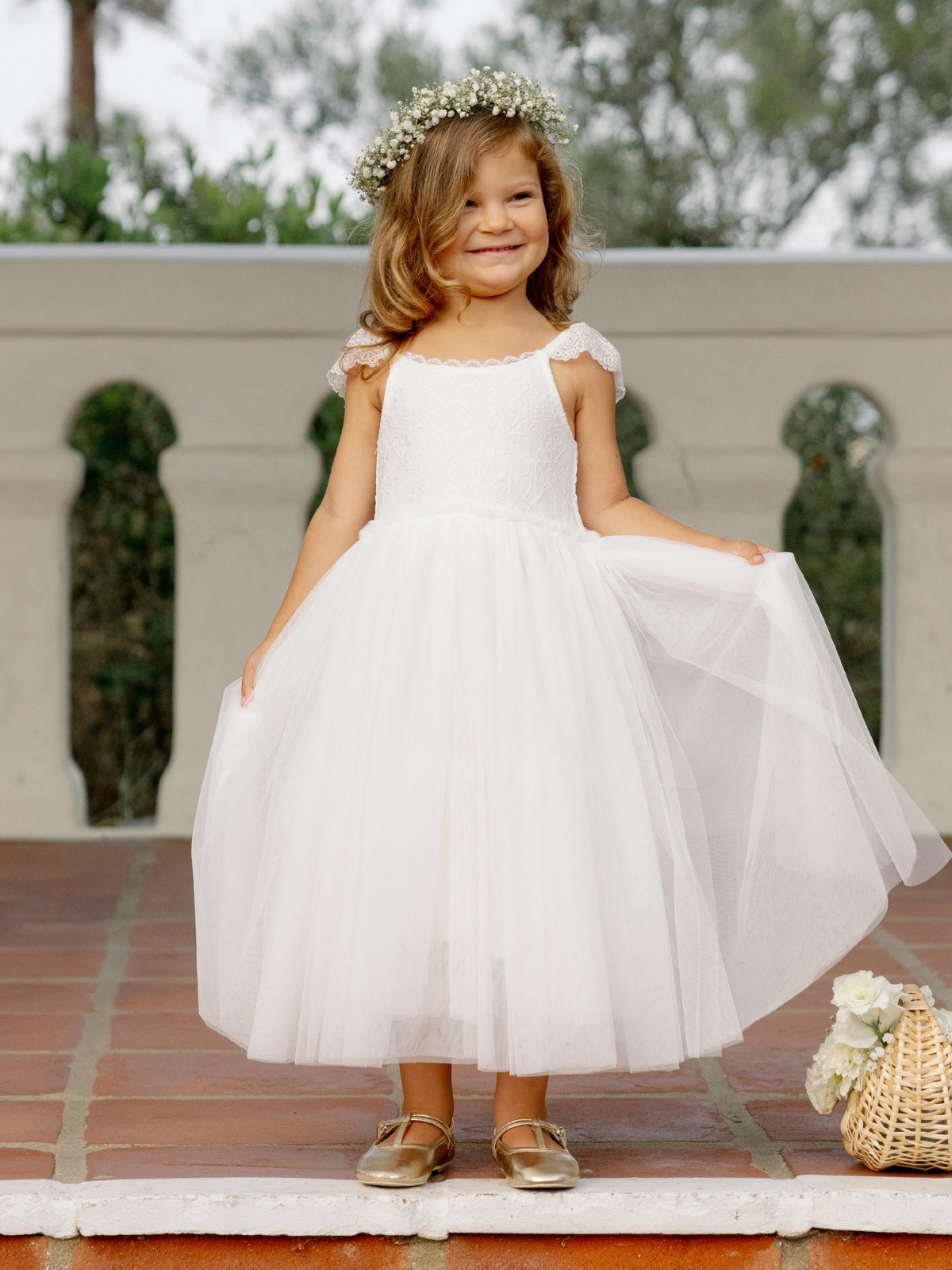 Smiling girl in a white dress and floral crown, standing on a patio beside a basket of flowers.