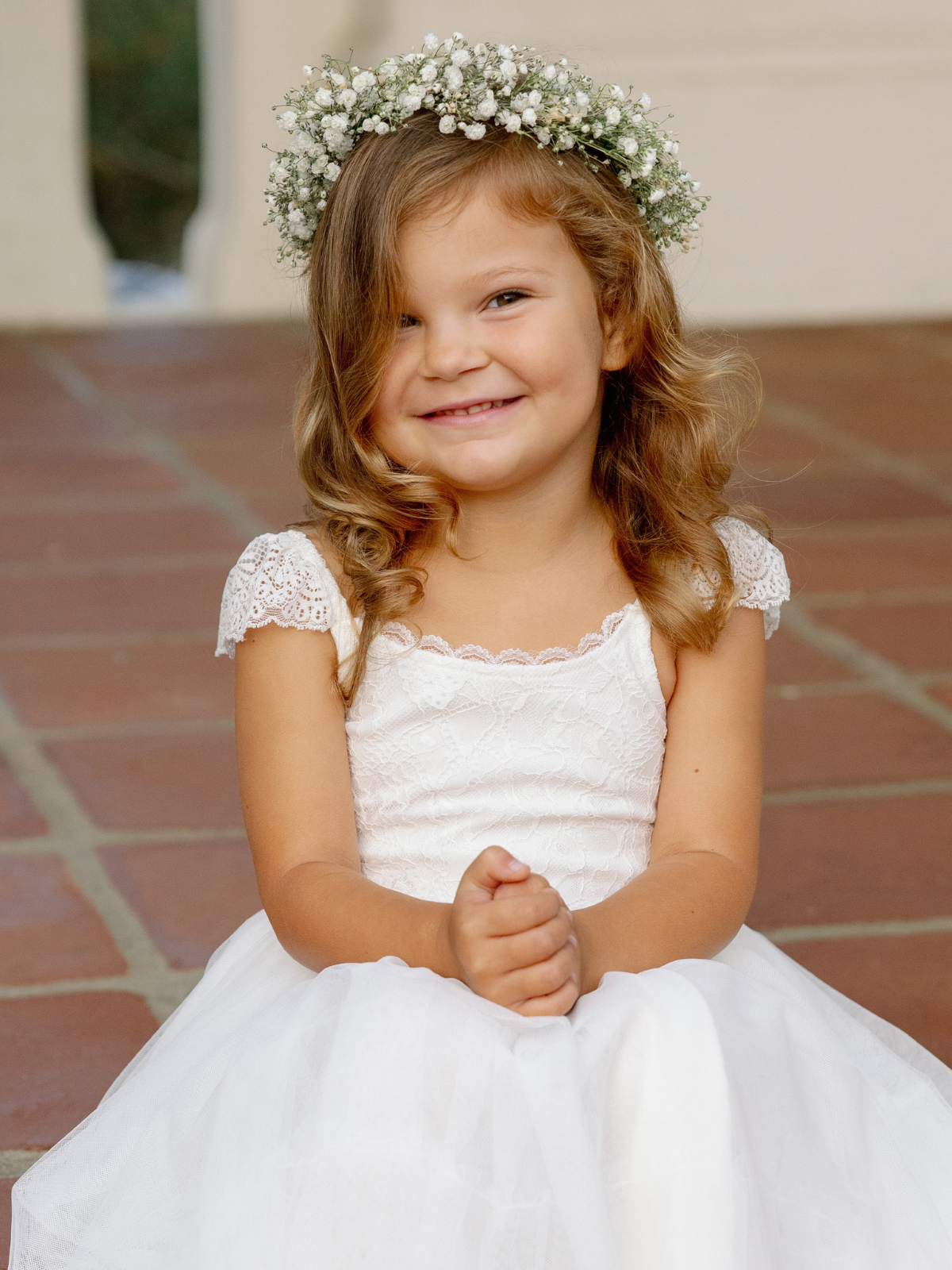 Smiling girl in a white dress and flower crown, sitting on a tiled surface.