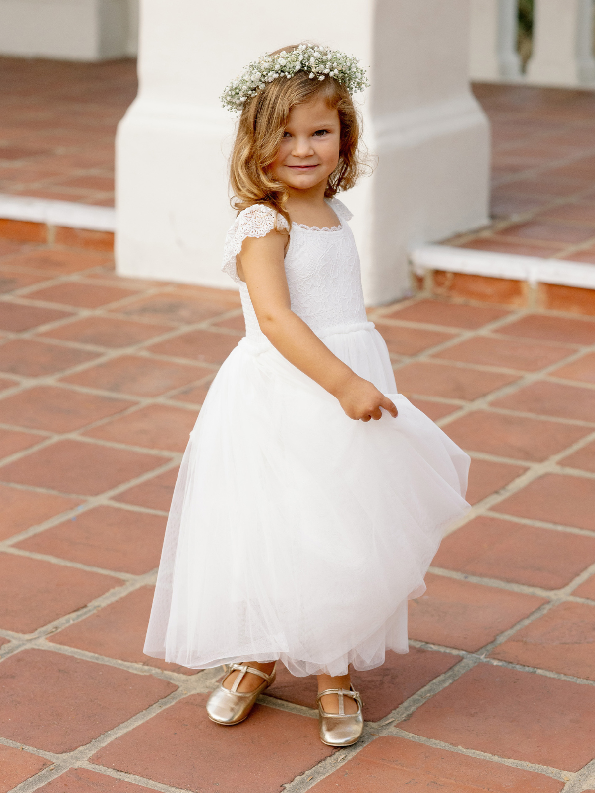 Smiling girl in a white dress and floral crown poses on tiled floor, showcasing her playful spirit.