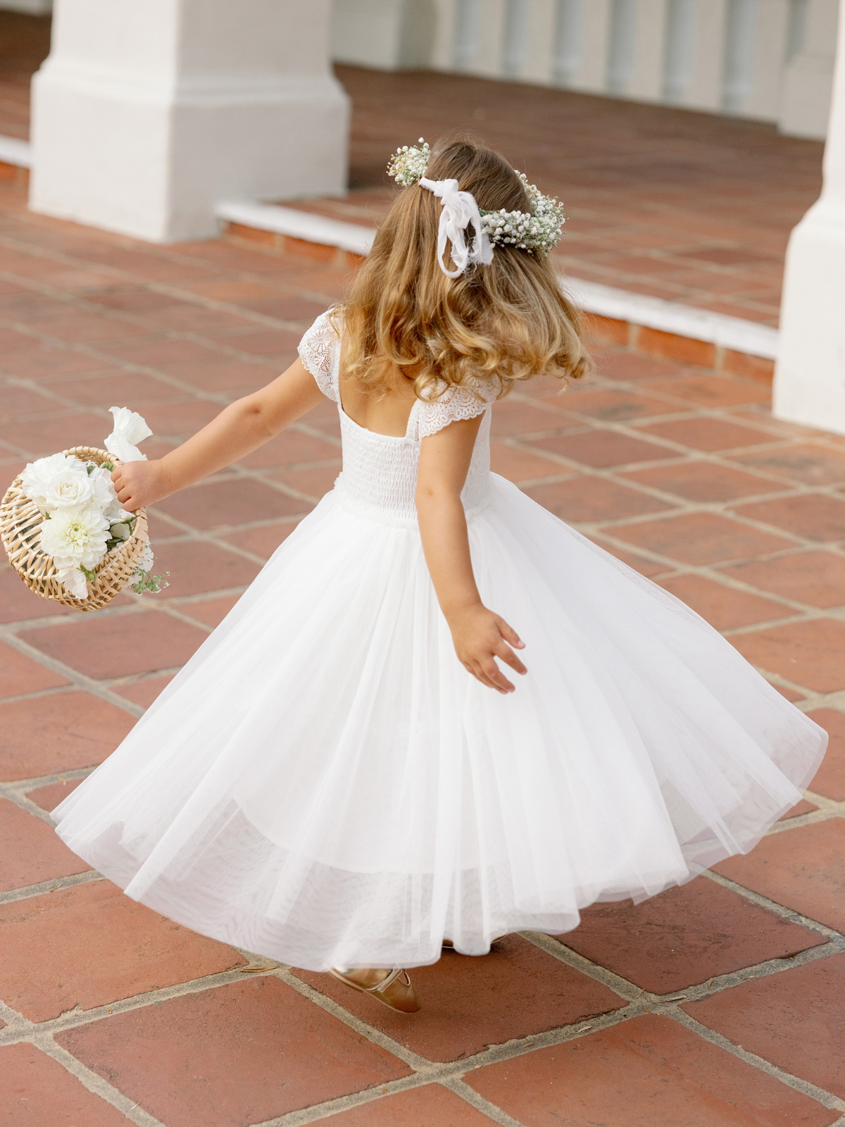Girl in a white dress twirls while holding a floral basket, with a charming outdoor patio in the background.