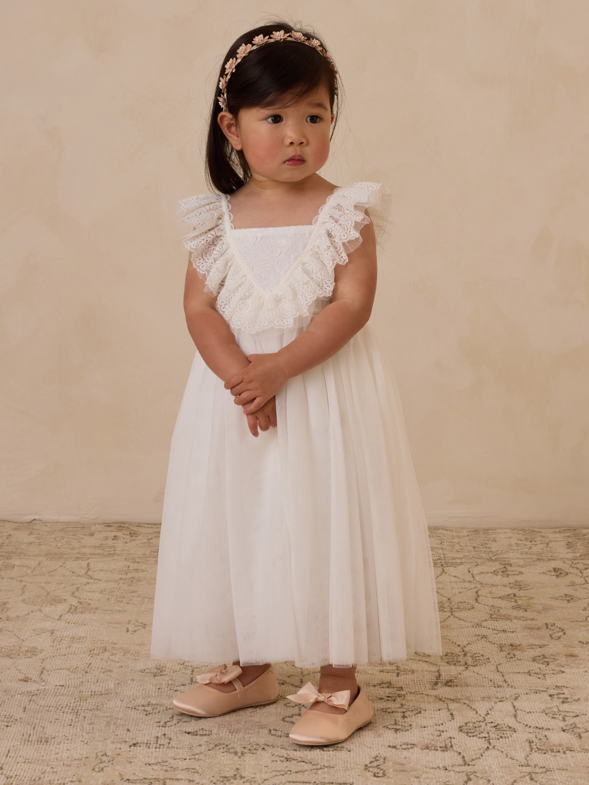 Young girl in a white dress with lace trim and floral headband, posing against a neutral background.