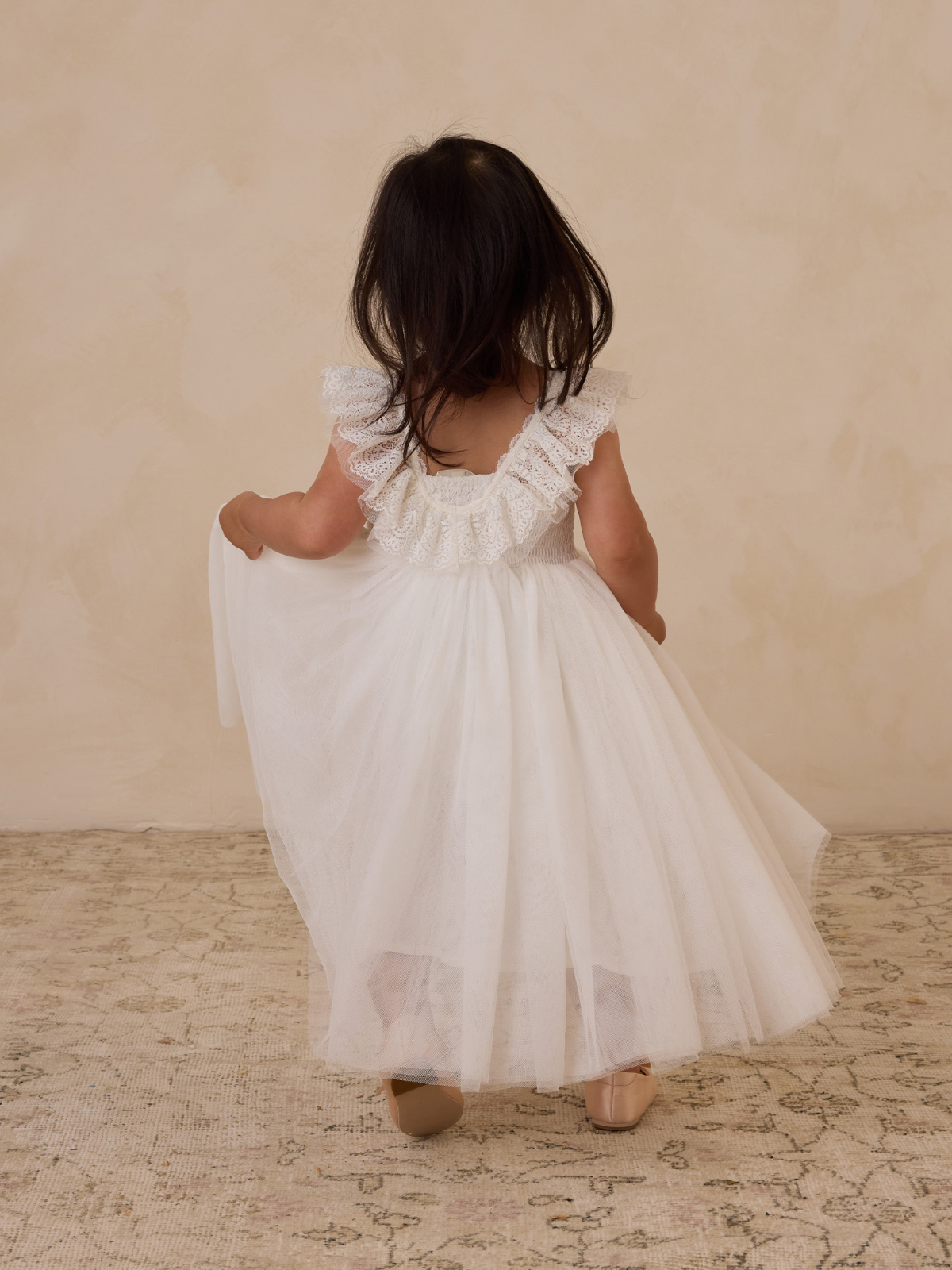 Toddler girl in a white dress with ruffles, facing away, on a textured rug against a soft beige backdrop.