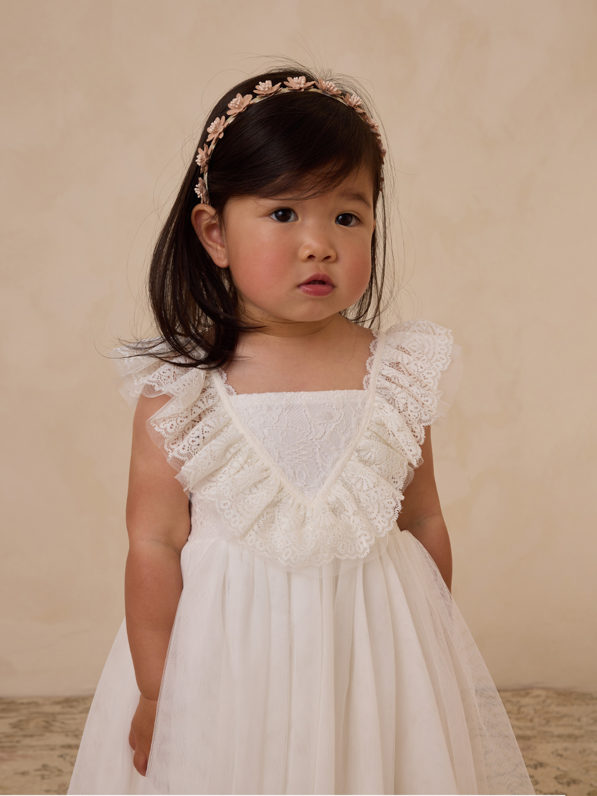 Child in a white lace dress with a ruffled neckline, wearing a floral headband, against a neutral background.