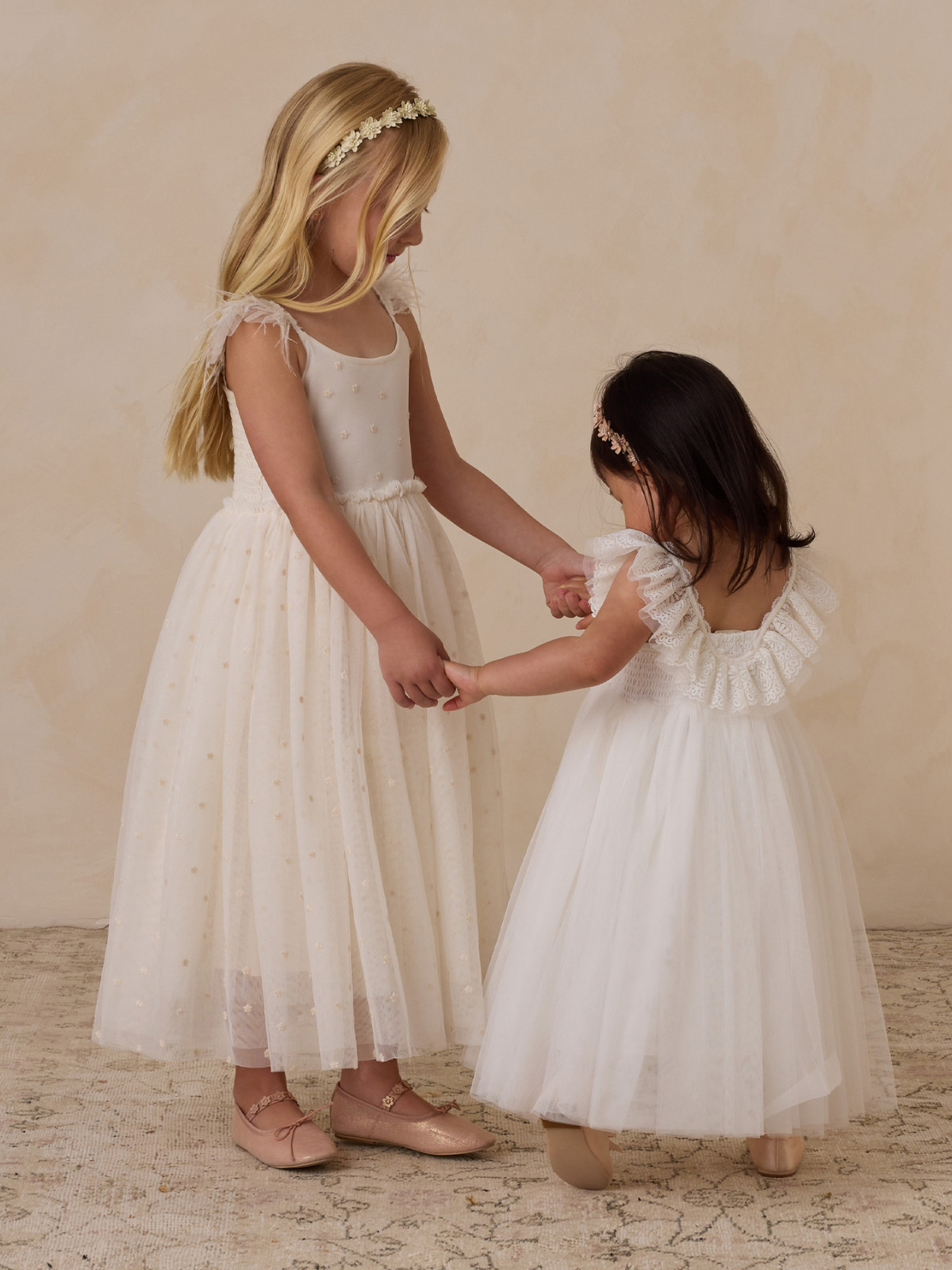 Two young girls in white dresses hold hands against a soft beige background, exuding joy and innocence.