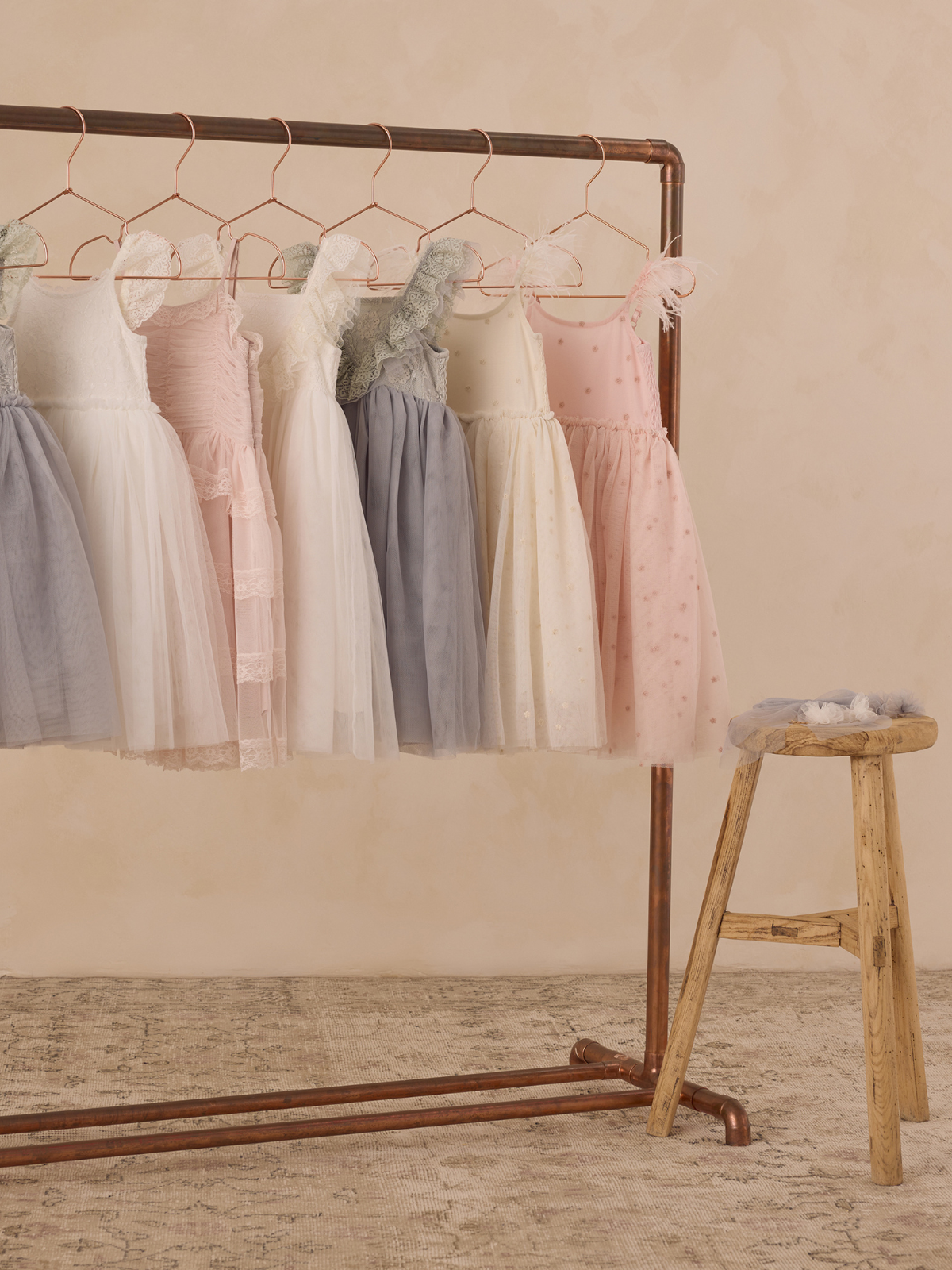 Assorted children's dresses hanging on a rose gold rack with a wooden stool nearby, set against a soft background.