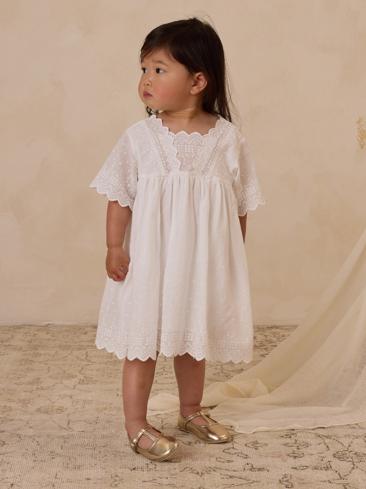 Young girl in a white dress with lace details, standing on a textured floor with a neutral background.