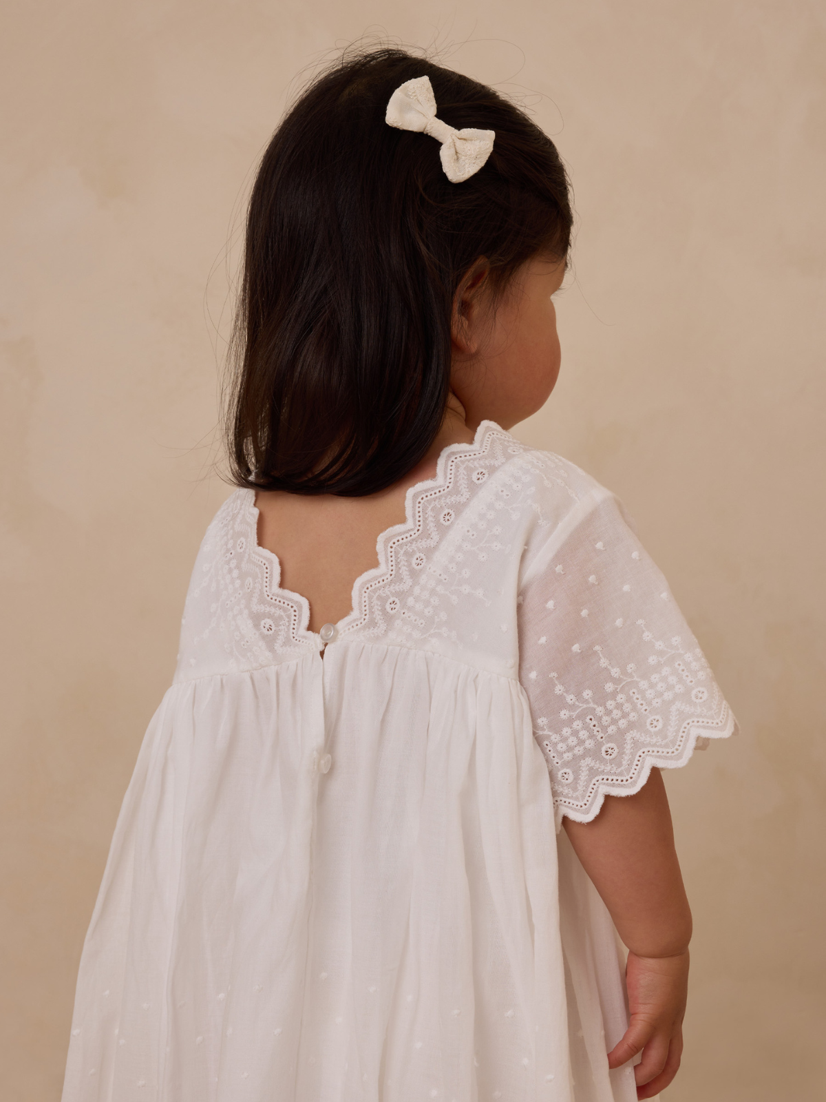 Toddler in a white embroidered dress with a bow in her hair, standing against a neutral background.