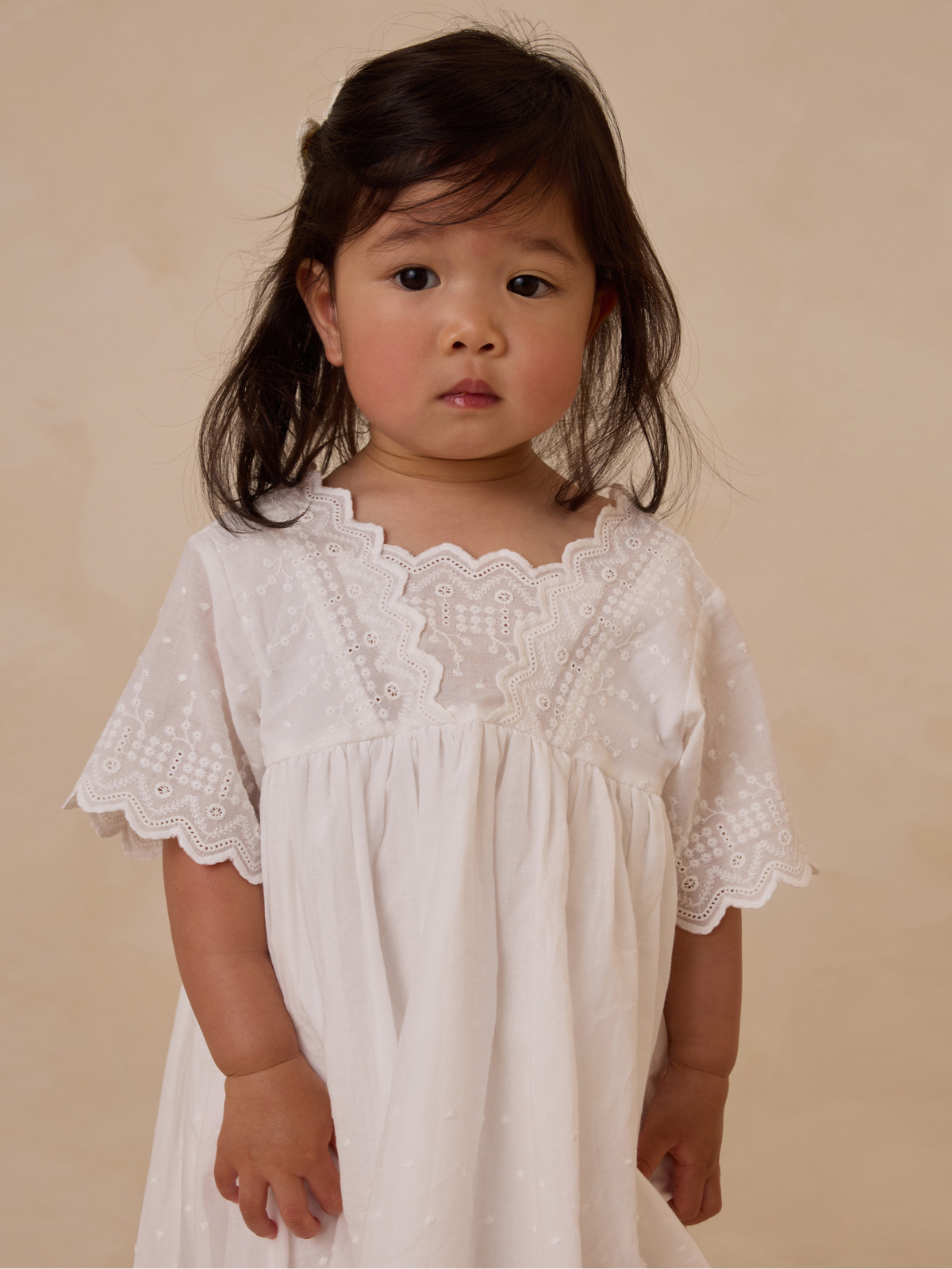 Child in a white dress with lace detailing, looking directly at the camera against a neutral background.