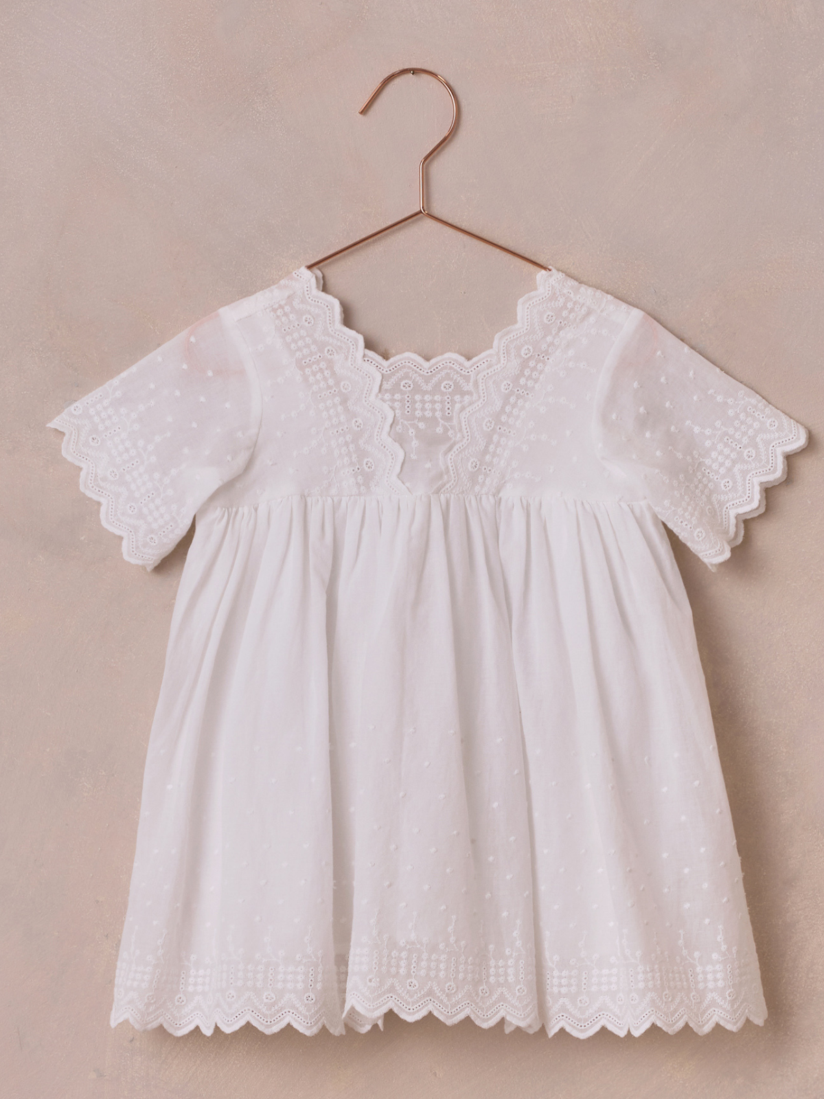 White children’s dress with lace detailing, hanging on a rose gold hook against a textured background.