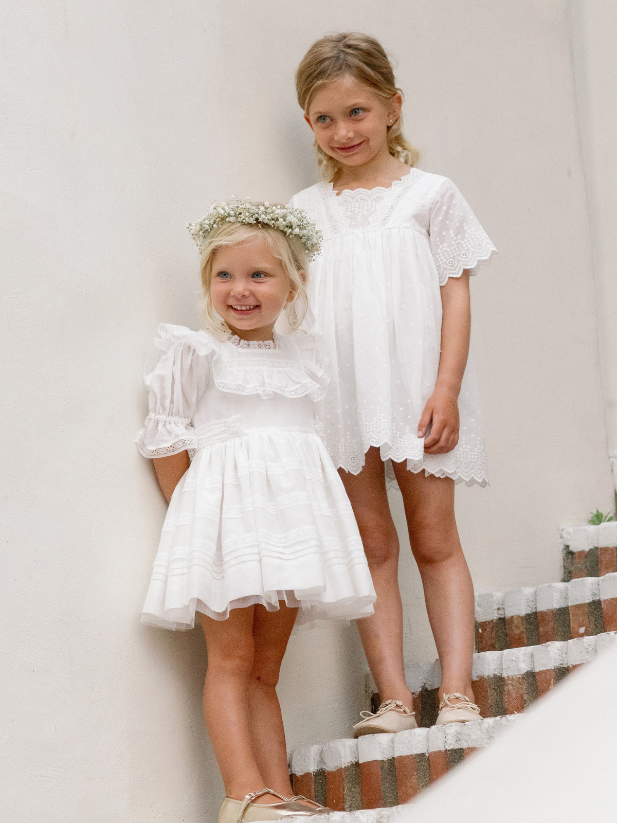 Two young girls in white dresses, one with a flower crown, standing on stairs against a light wall.