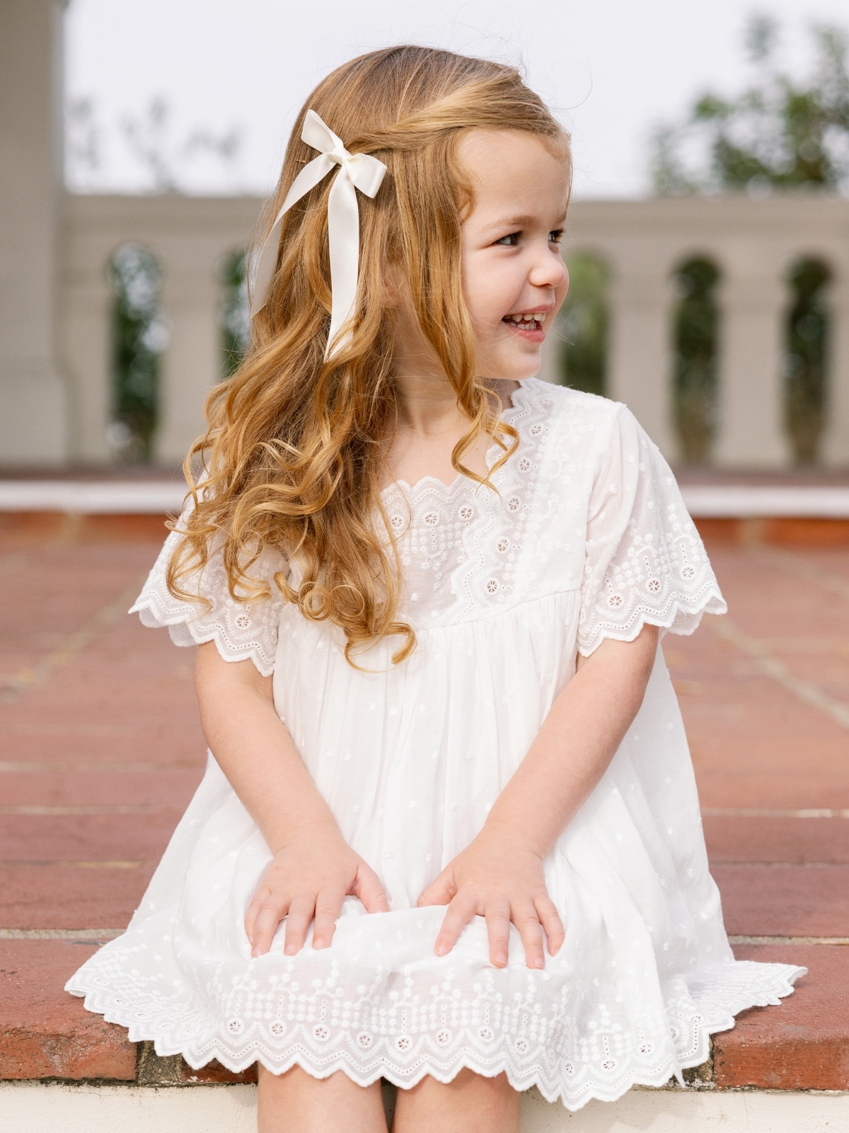 Smiling girl with long curly hair in a white dress sitting on steps, adorned with a ribbon in her hair.
