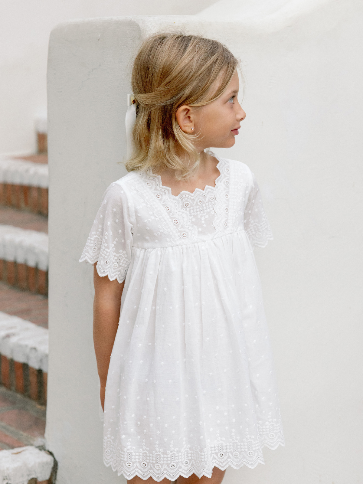 A young girl in a white dress with lace detail stands against a light background, looking to the side.
