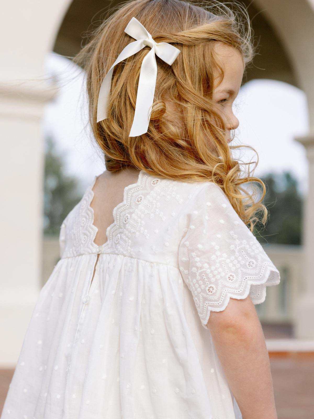 Young girl with curly hair and a bow wearing a white embroidered dress, standing outdoors in soft light.