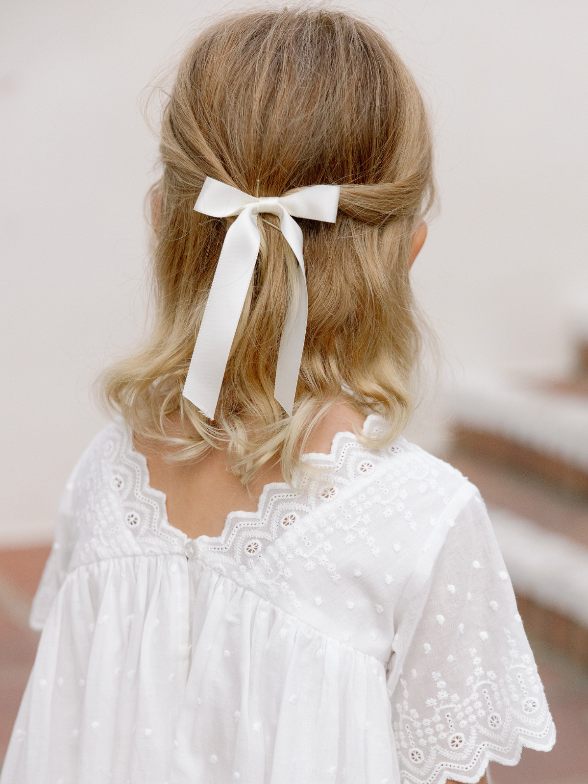 Girl with wavy hair wearing a white dress and a bow, viewed from the back. Soft, natural setting.