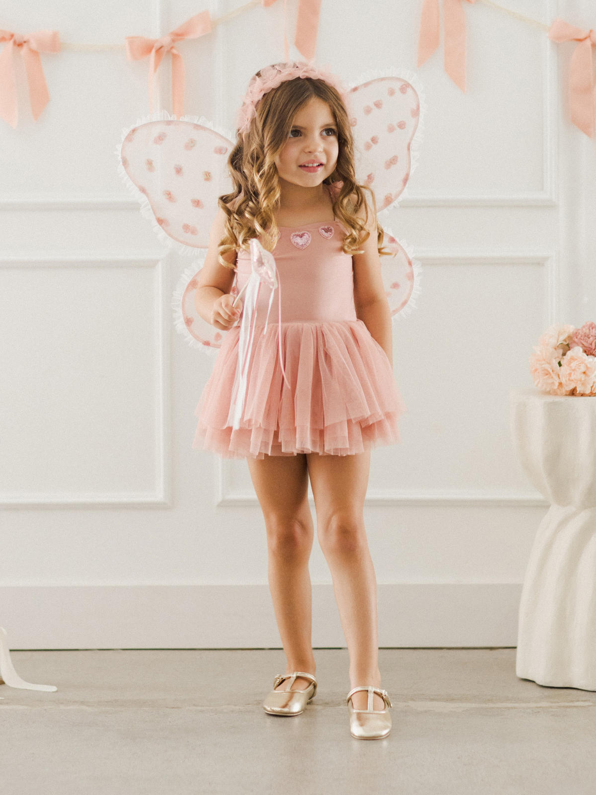 Girl in a pink fairy costume with wings, holding a wand, standing in a decorated room.
