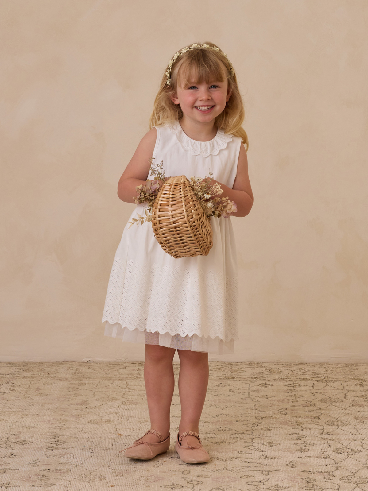 Smiling girl in a white dress holding a woven basket with flowers, against a soft beige background.