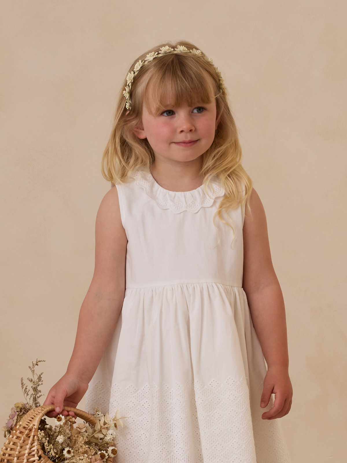 Young girl in a white dress holding a basket of flowers, smiling softly against a neutral background.