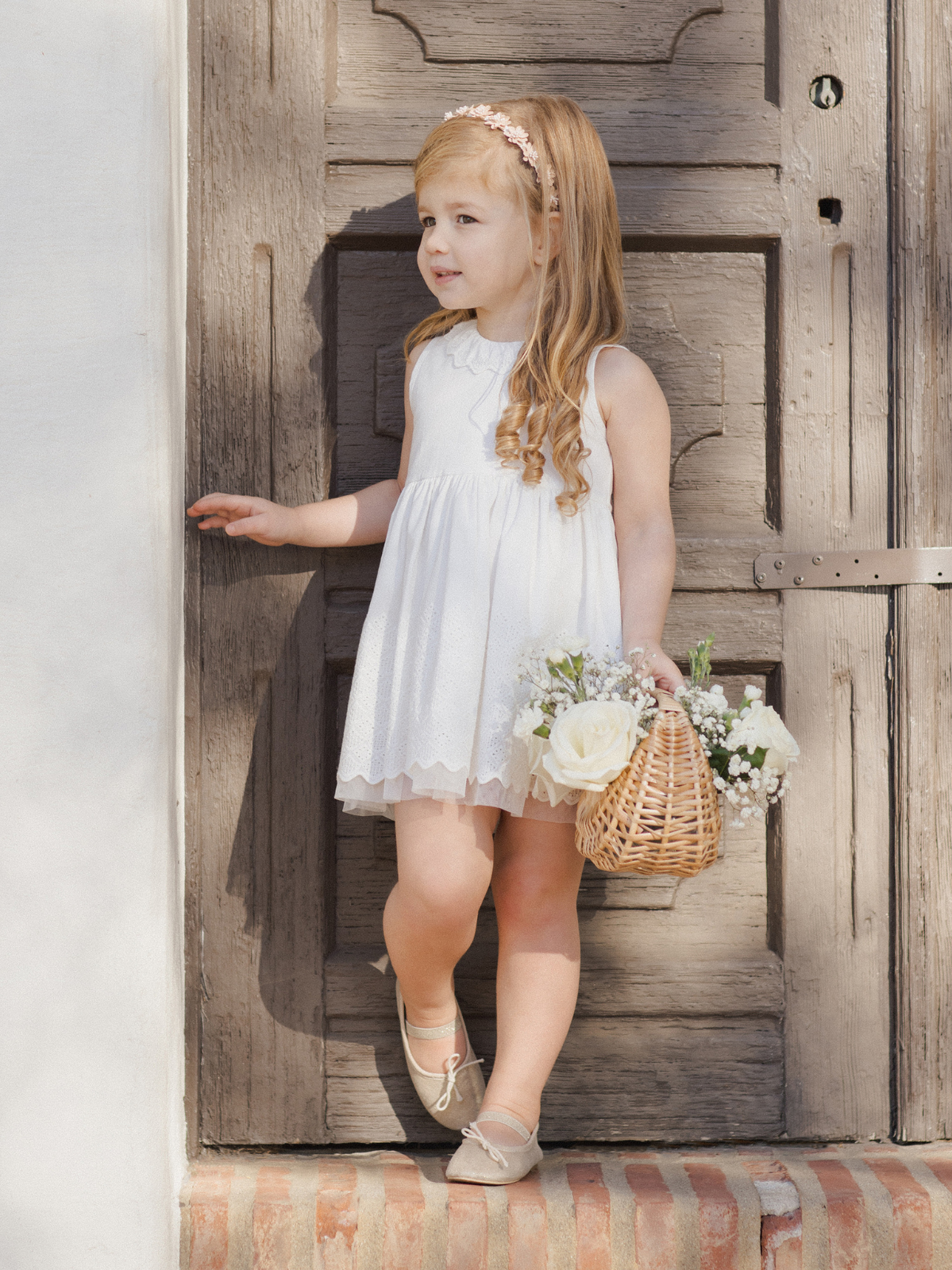 Girl in a white dress holding a flower basket, standing by a wooden door. Soft sunlight highlights her smile.